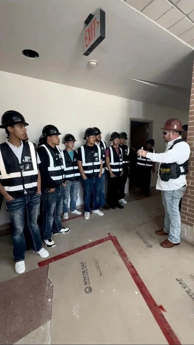 A construction safety training session with students wearing helmets and safety vests, listening to an instructor gesturing with his hands indoors near a brick wall and an exit sign.