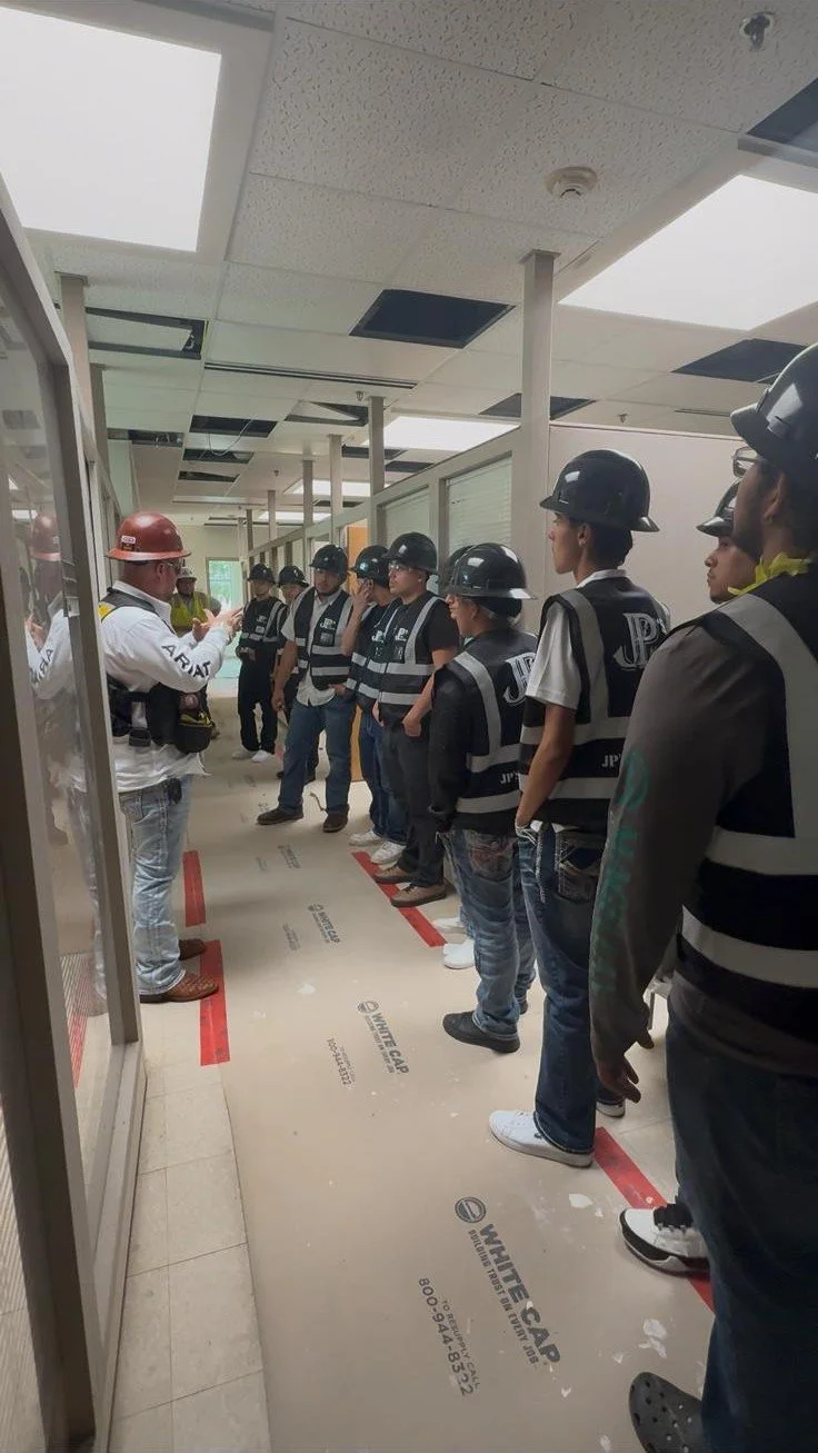 A group of people wearing construction helmets and vests standing in a hallway, listening to a person with a red helmet who appears to be giving instructions or a briefing.