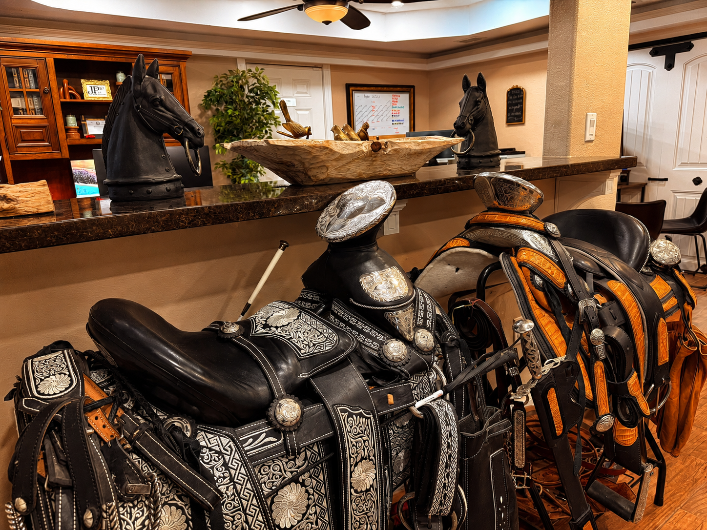 Indoor room with two decorative horse head sculptures on a dark brown marble counter, two ornate cowboy saddles with intricate patterns and silver embellishments, and a wooden bowl with decorative birds on the counter. In the background, there's a green plant, a framed whiteboard, and a black ceiling fan.