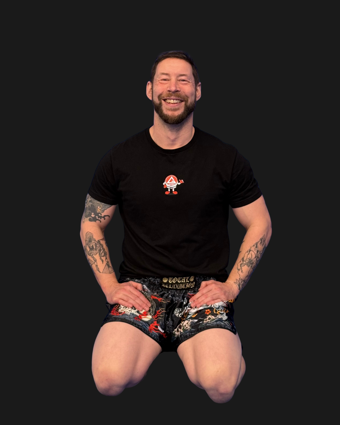 A man kneeling down, smiling, wearing a black t-shirt with a cartoon character and martial arts shorts with a dragon design, against a black background, with text on the left listing kickboxing and Muay Thai.