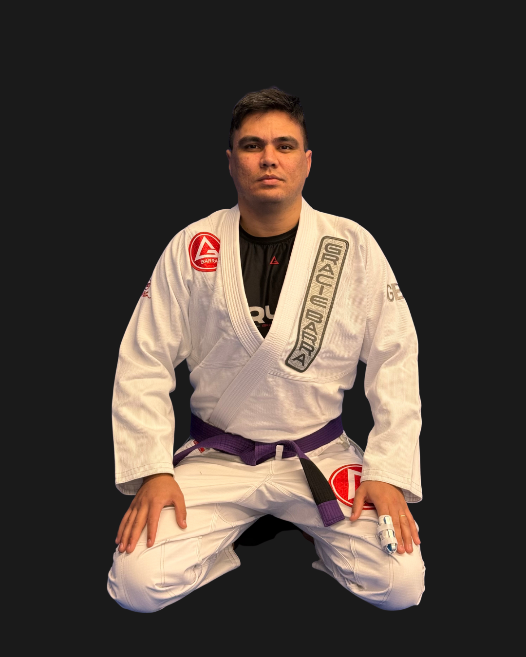A man in a white Brazilian Jiu-Jitsu gi with patches and a purple belt, kneeling on the ground with a serious expression, against a plain dark background.