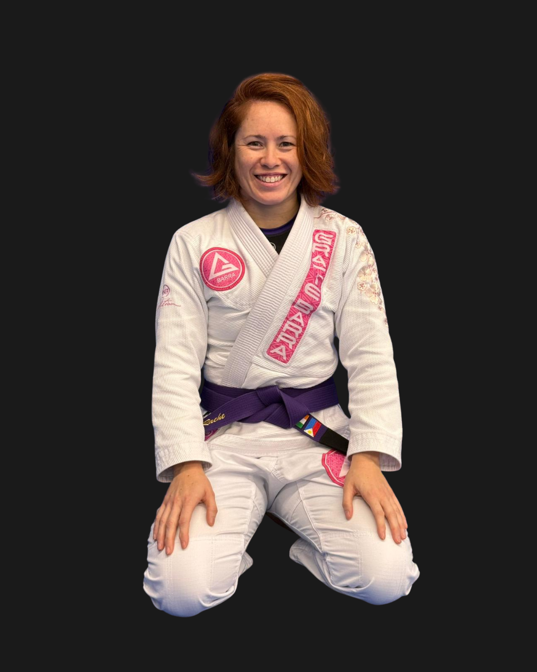 Female Brazilian Jiu-Jitsu athlete wearing a white gi with pink patches, sitting on her knees on a black background. She has red hair and is smiling.