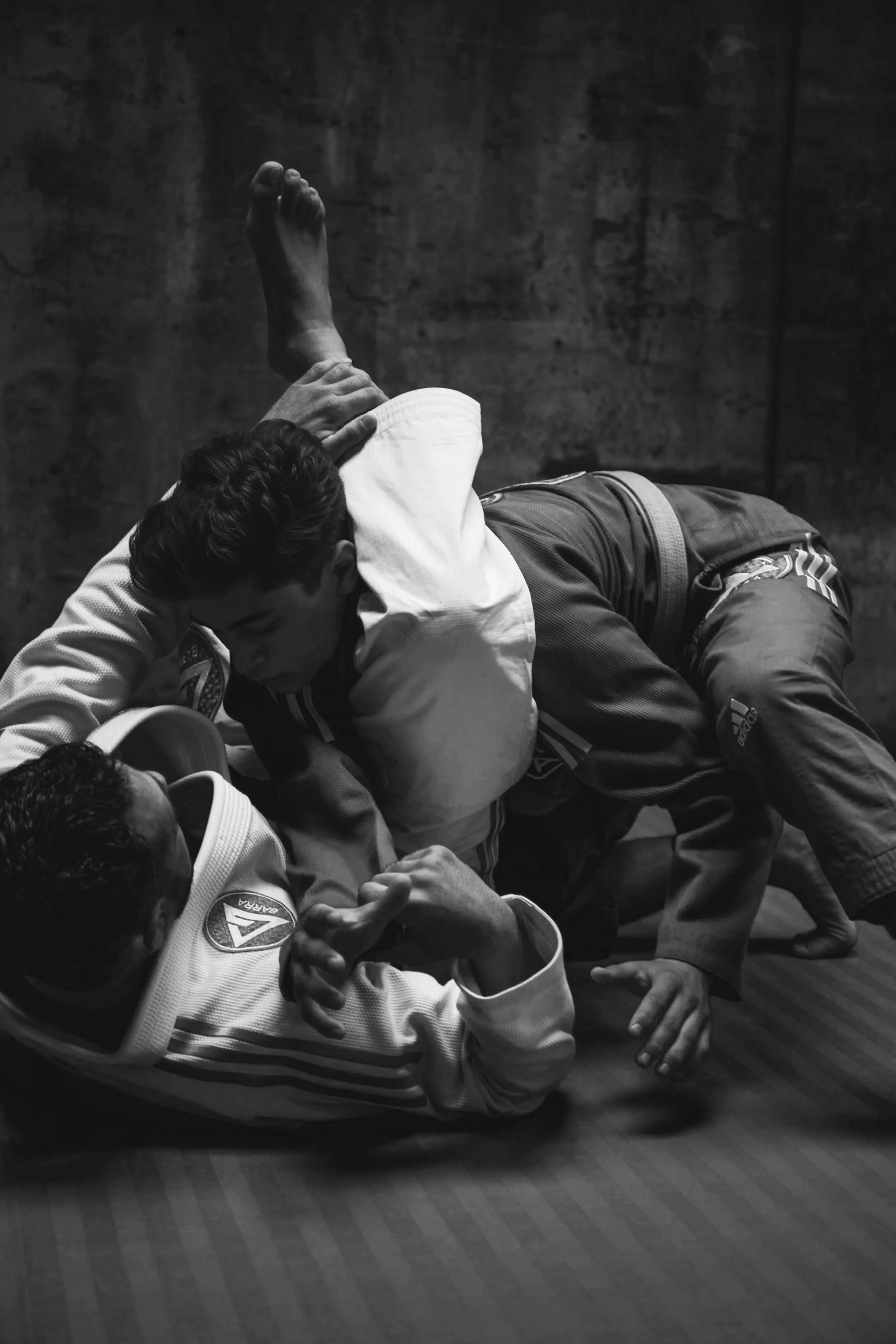 Two martial artists practicing Brazilian Jiu-Jitsu on a mat, with one in a white gi and the other in a dark gi, engaged in a ground fighting position.