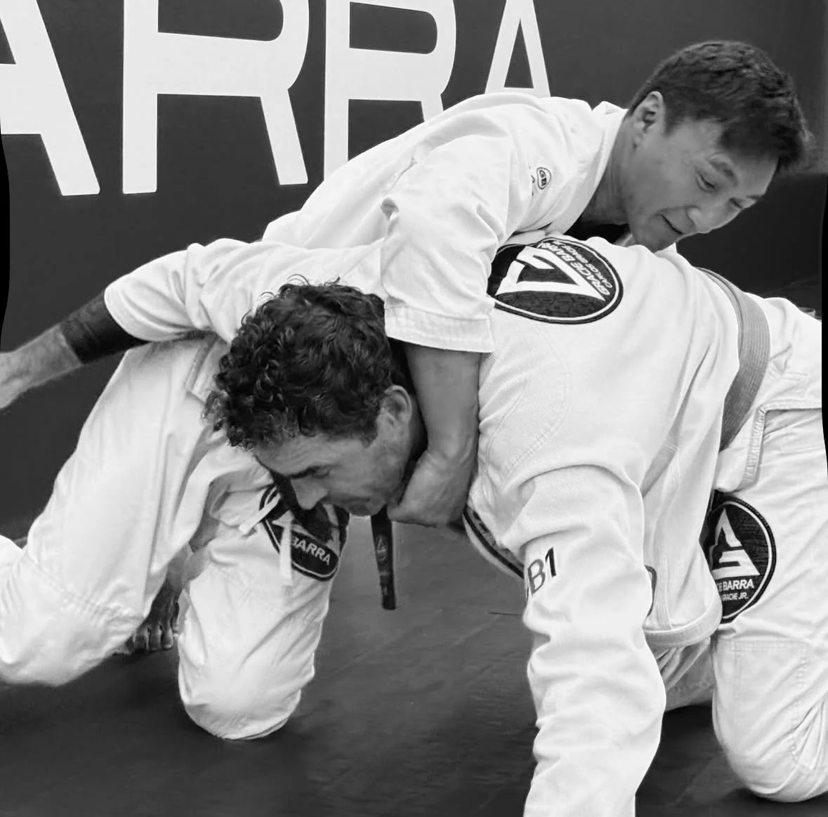 Two martial artists practicing Brazilian Jiu-Jitsu on the mat, one in a white gi on top, the other in a white gi underneath, with a large logo on the back of the top practitioner's gi.