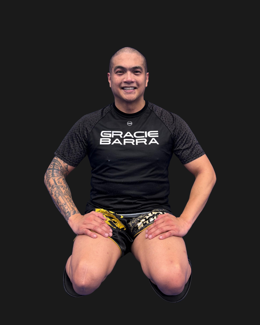 A man with short hair and tattoos on his left arm, wearing a black rash guard with the words 'GRACIE BARRA' printed on it and black and yellow shorts, kneeling with a smile against a plain black background.