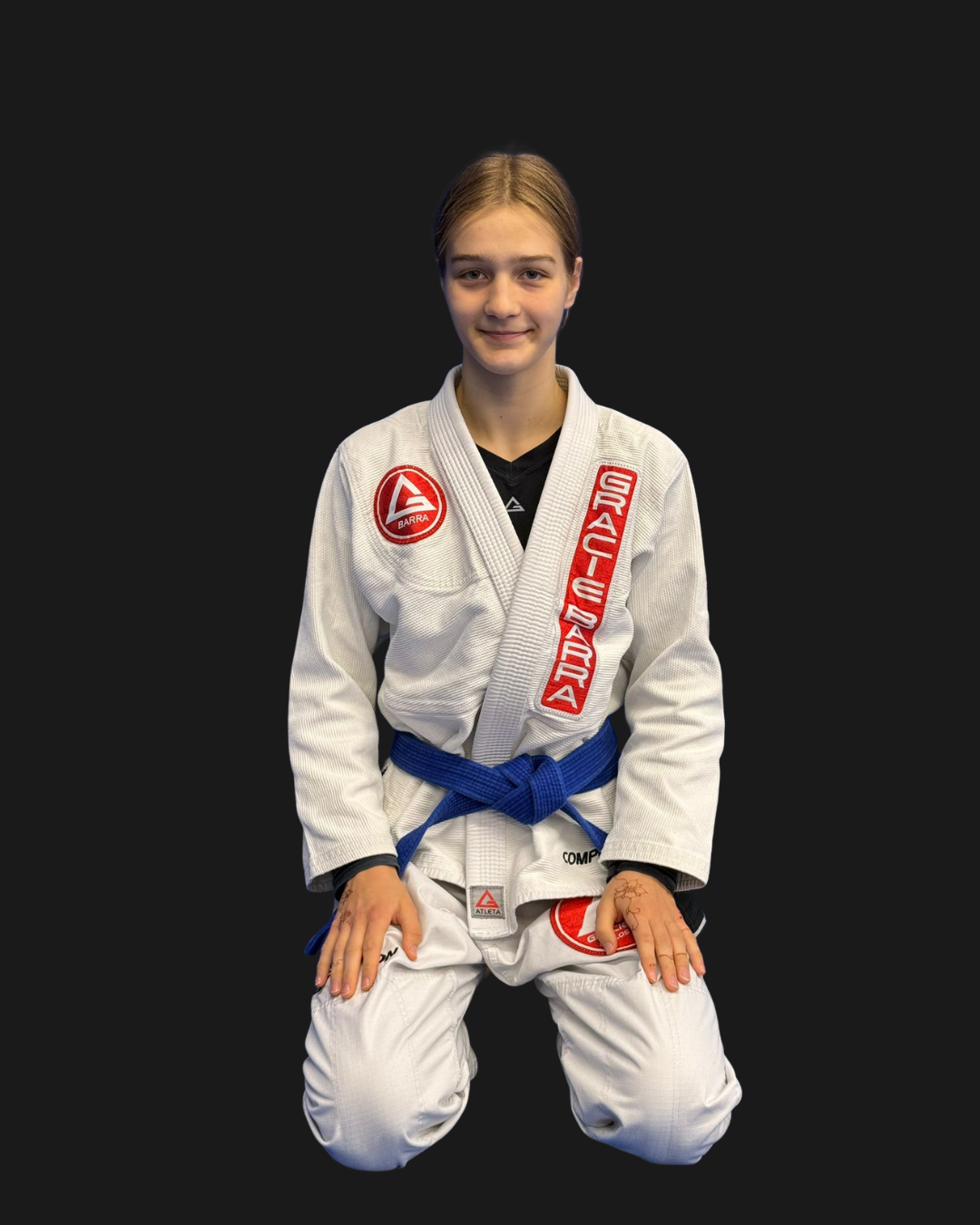 Young woman in a white Brazilian Jiu-Jitsu gi with red and blue patches, kneeling on the ground, smiling for the camera against a black background.