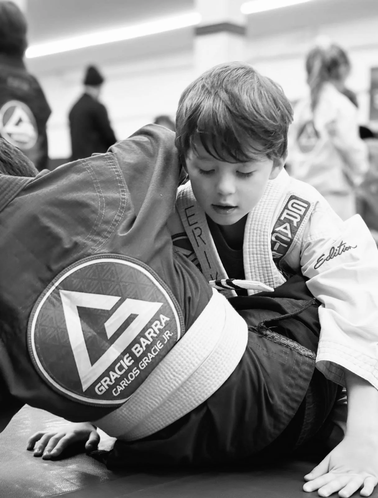 A young boy practicing Brazilian Jiu-Jitsu on an adult training partner, who is wearing a gi with a patch that reads 'Grace Barra.' The boy appears focused, and the background shows other practitioners and instructors in a martial arts training facility.