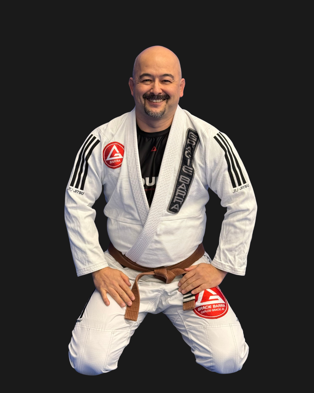 Man in a blue Brazilian Jiu-Jitsu gi with brown belt, kneeling on the mat, smiling, with patches and text on his gi, against a black background.