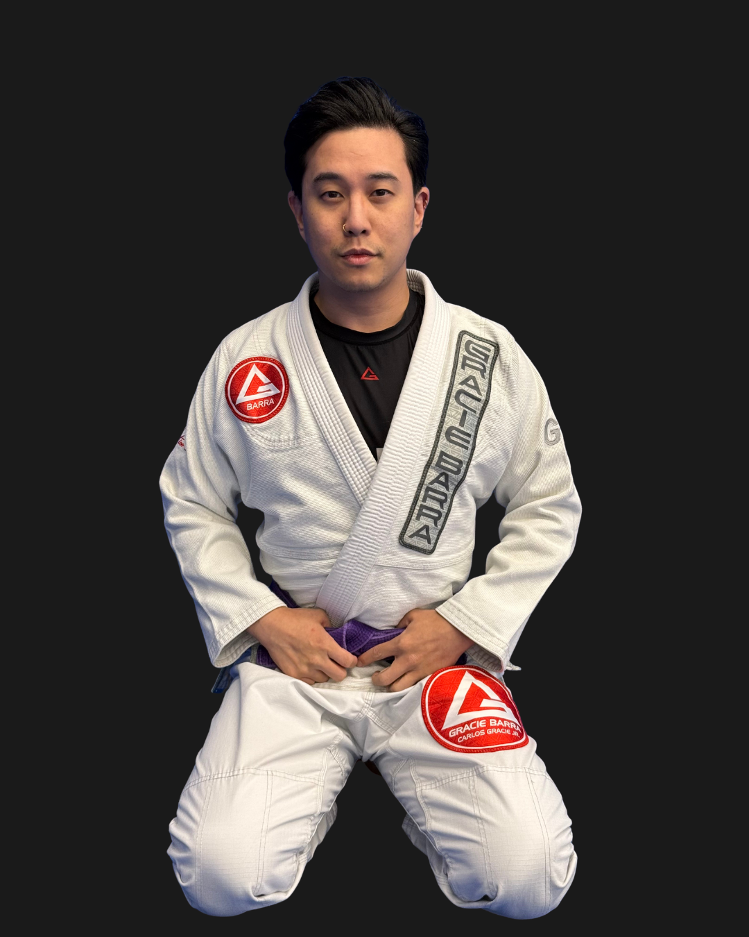 A male Brazilian Jiu-Jitsu practitioner wearing a white gi with patches and a black rash guard underneath, kneeling against a black background, posing confidently.