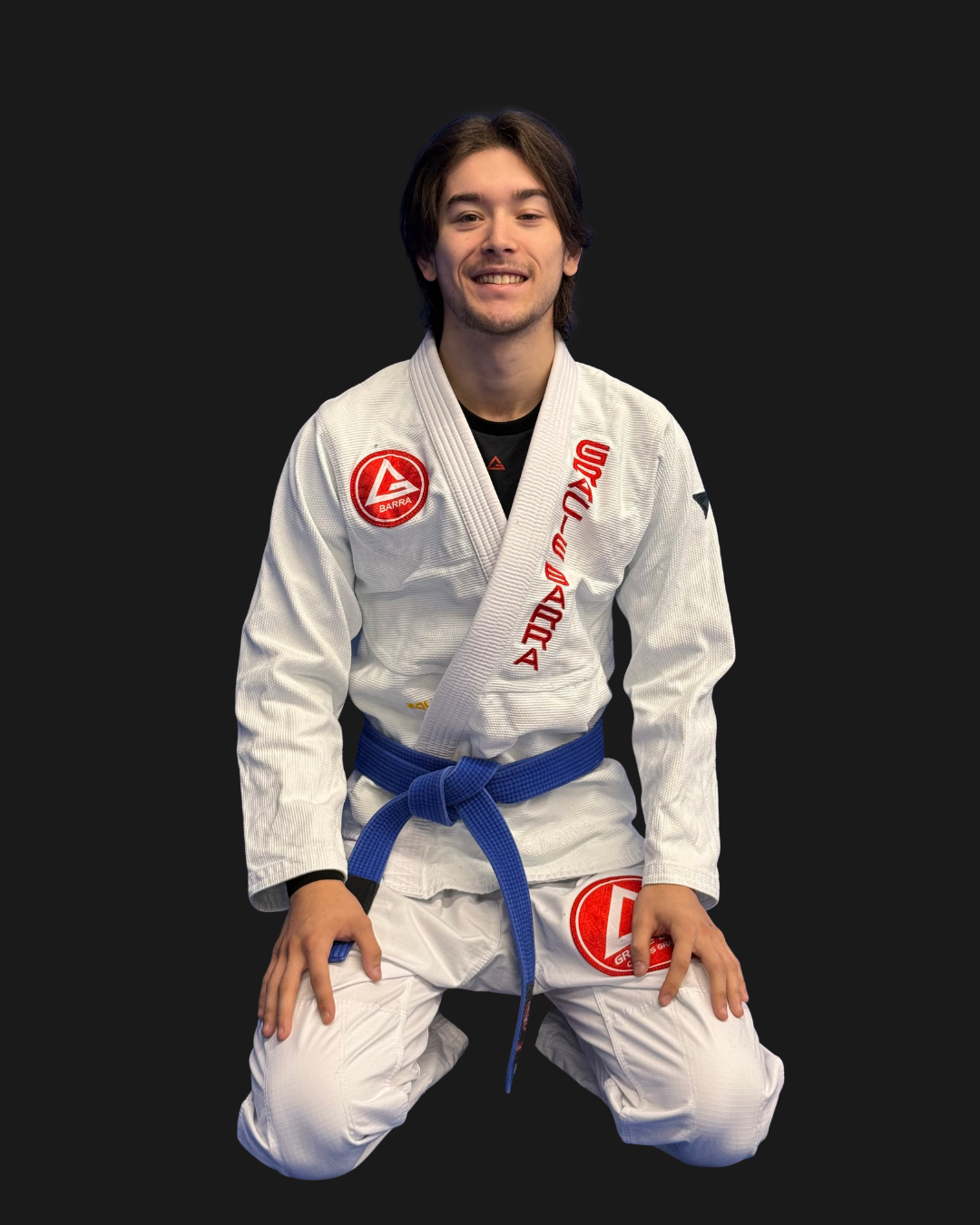 A young man in a white Brazilian Jiu-Jitsu gi with red patches and a blue belt, kneeling on the floor with a dark background. Text on the image reads 'Coach Hiro' and 'GB' with a green 'K'.