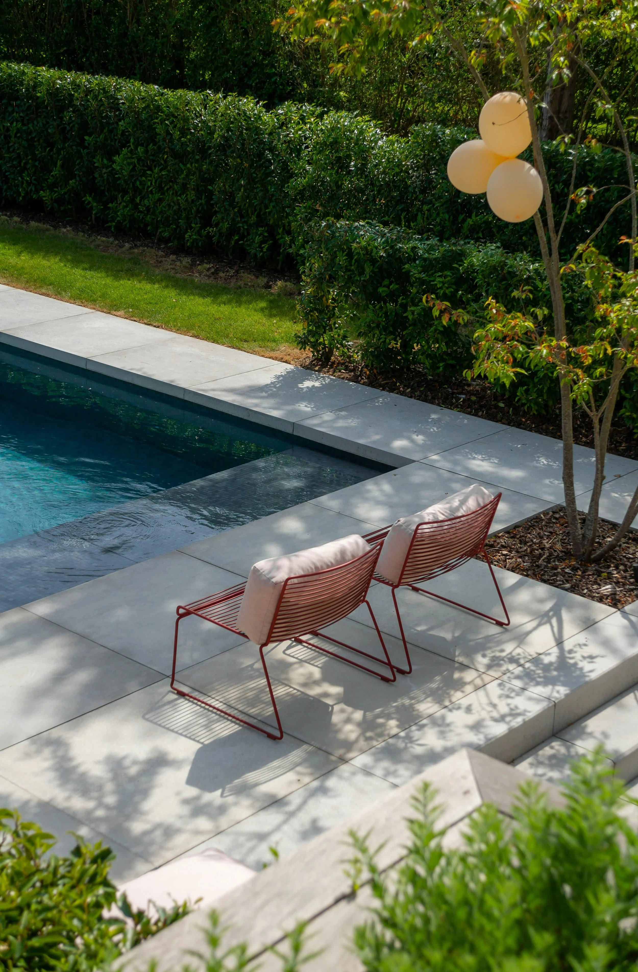 Dos sillas de metal rojas con cojines blancos junto a una piscina, árboles y arbustos verdes, y globos beige amarrados a un árbol