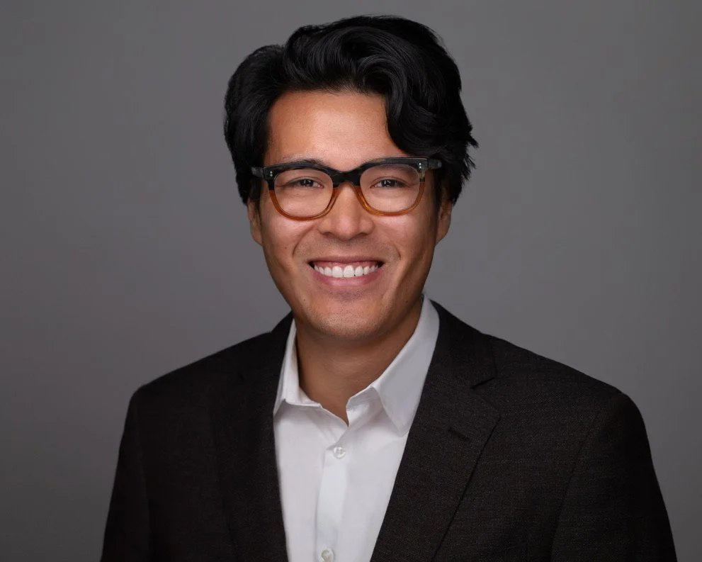 A young man with dark hair, glasses, and a broad smile wearing a black suit jacket and white shirt against a gray background.