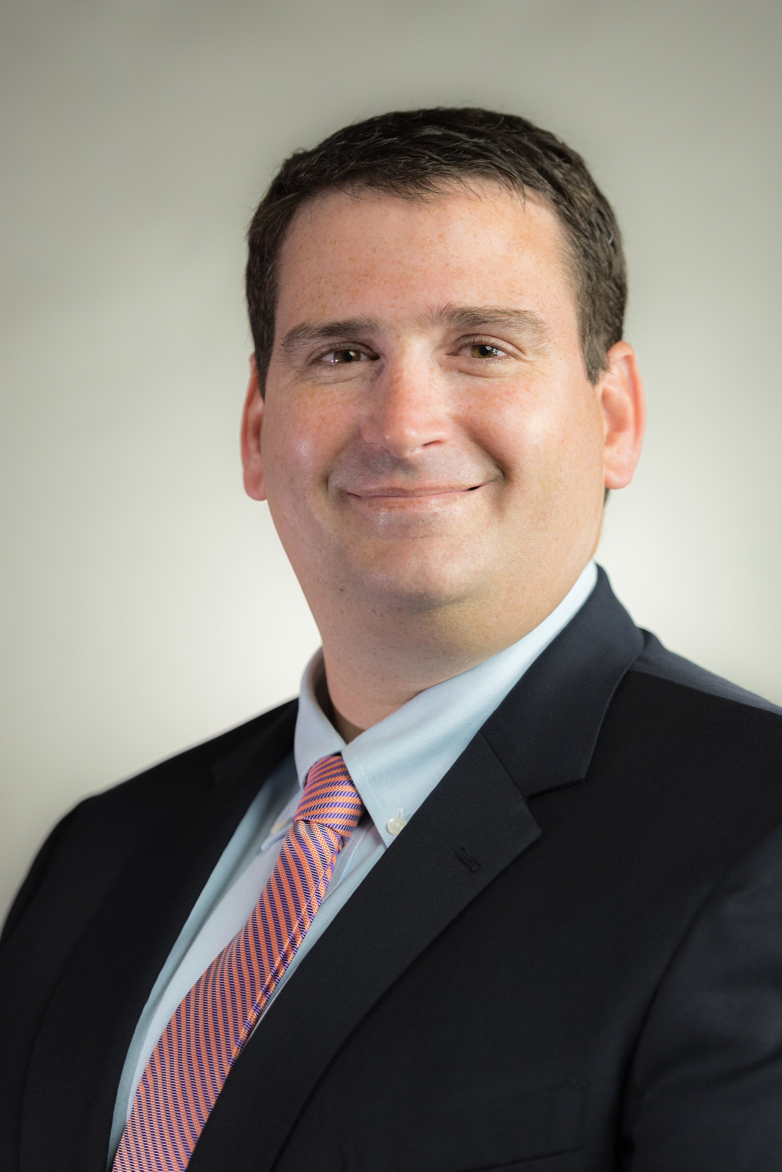 Professional headshot of a man in a suit with a pink and purple striped tie, smiling lightly, against a plain background.
