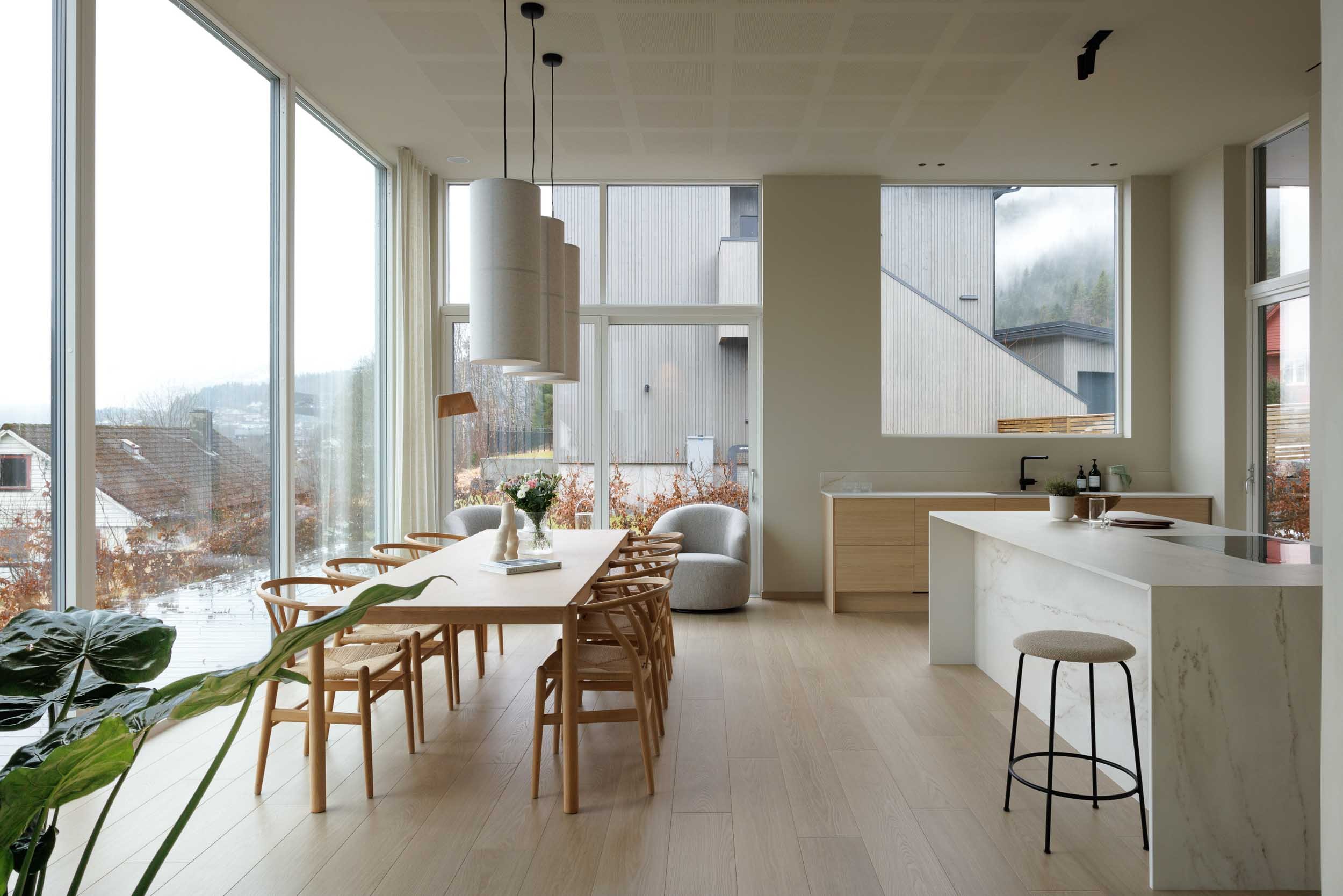 Modern dining area with large floor-to-ceiling windows, wooden dining table with matching chairs, a gray armchair, hanging pendant lights, and a kitchen island with a stool, all in a neutral color palette. - Førde, Sunnfjord