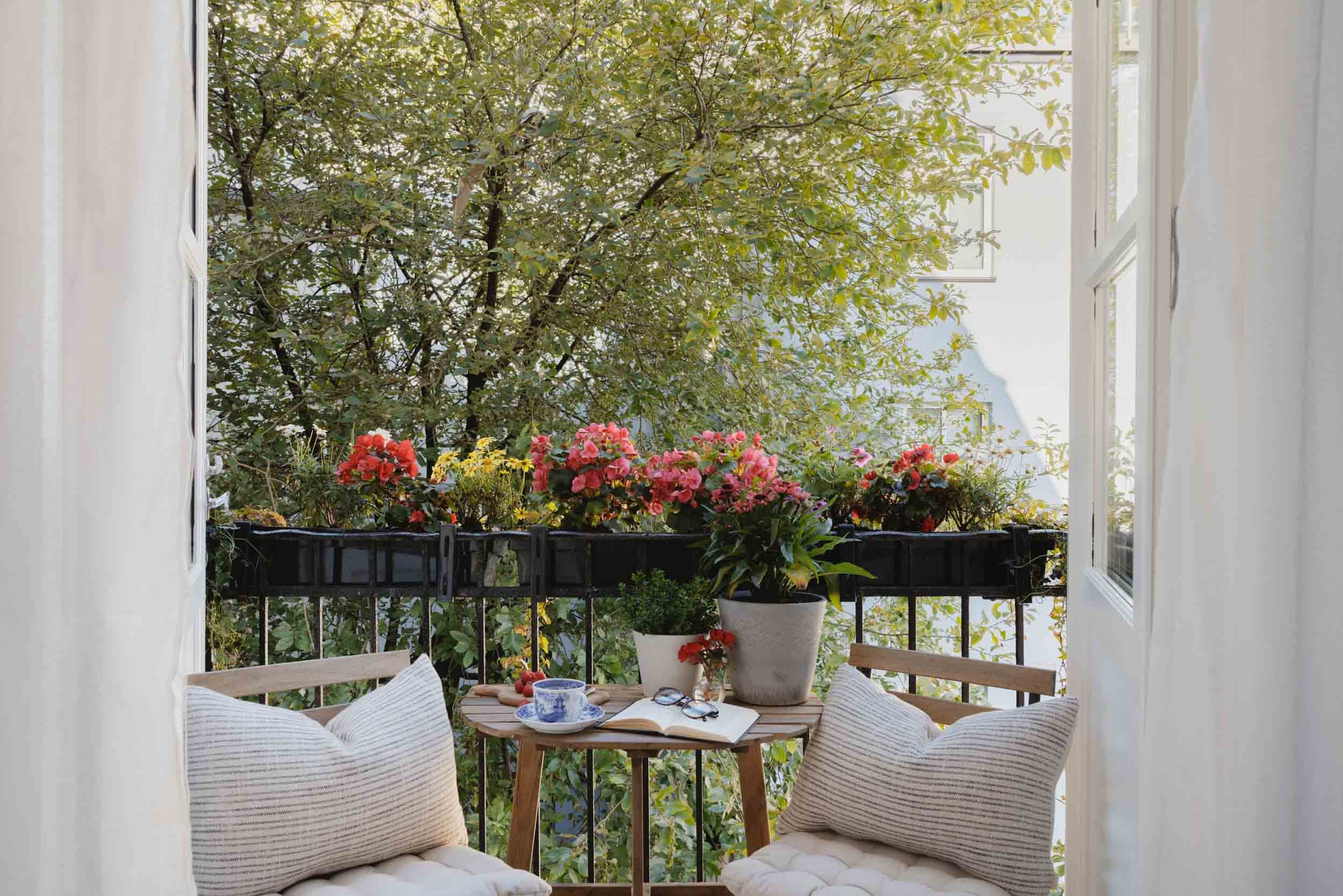 Cozy balcony with a small round wooden table, two cushioned chairs, potted plants, blooming flowers, and view of trees outside framed by open doors