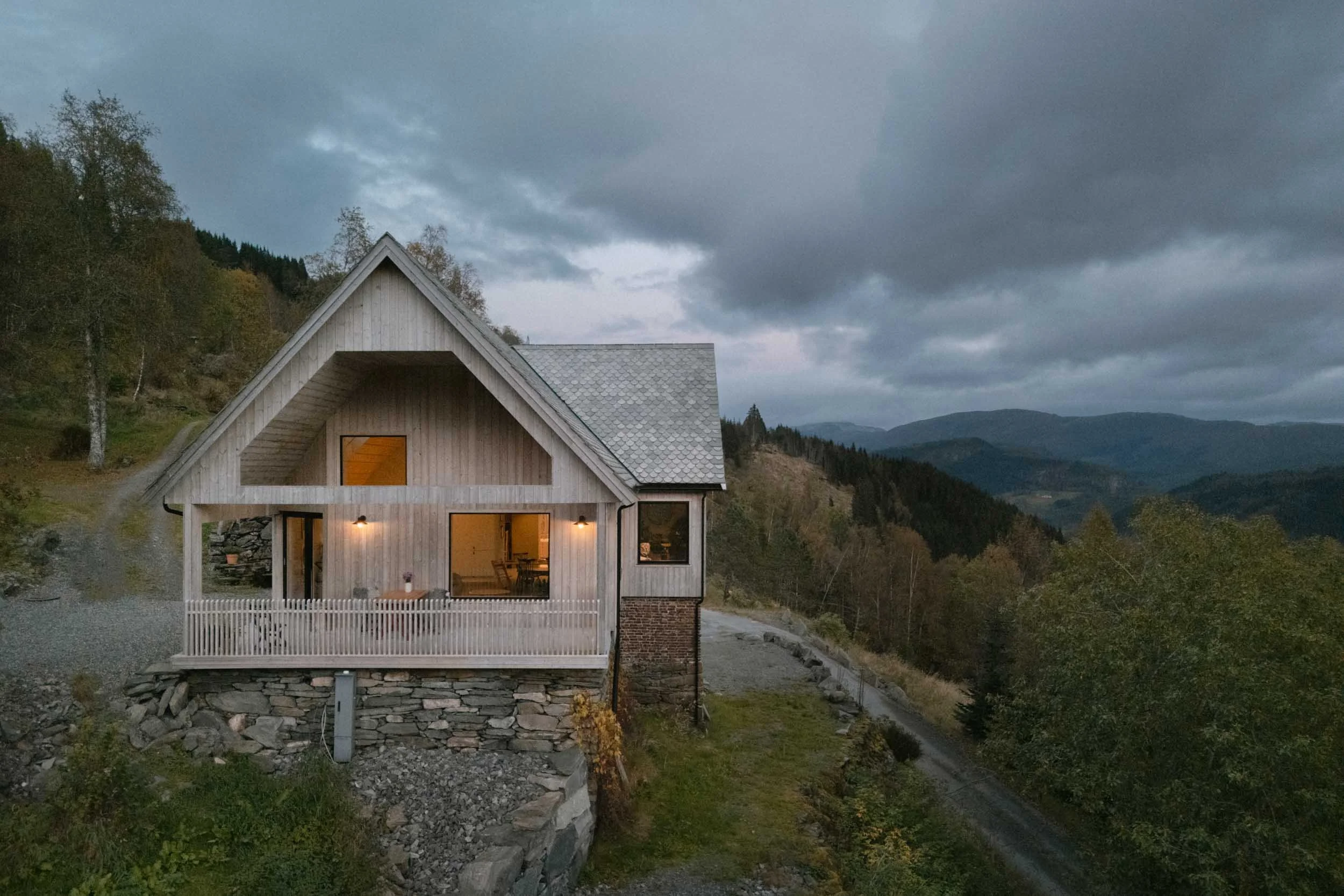 A rustic house on a hillside surrounded by trees and mountains under a cloudy sky at dusk.
