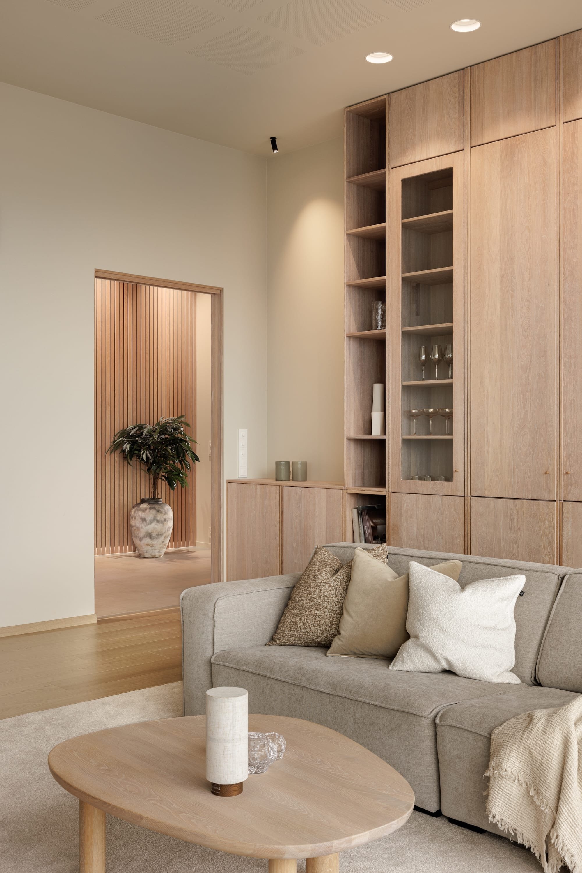 A living room with a beige sofa with pillows, a wooden coffee table, and a built-in wooden cabinet with open shelves and closed storage. There is a doorway leading to a space with a wooden slatted wall and a large potted plant.