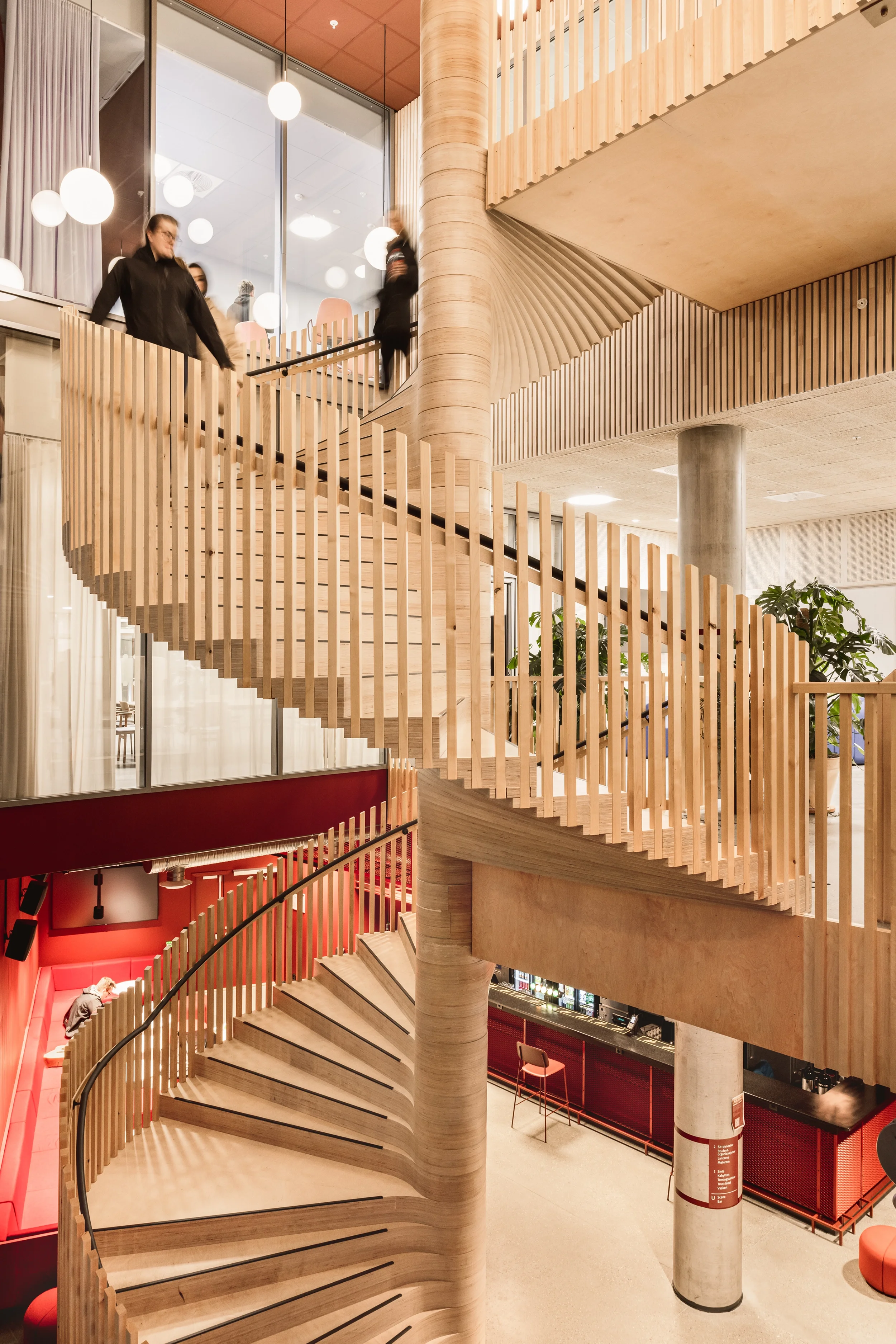 Indoor space featuring a modern, curving wooden staircase with vertical slats, connecting multiple floors with glass railings and a cozy seating area with red walls and furniture. - Student building University of Ålesund