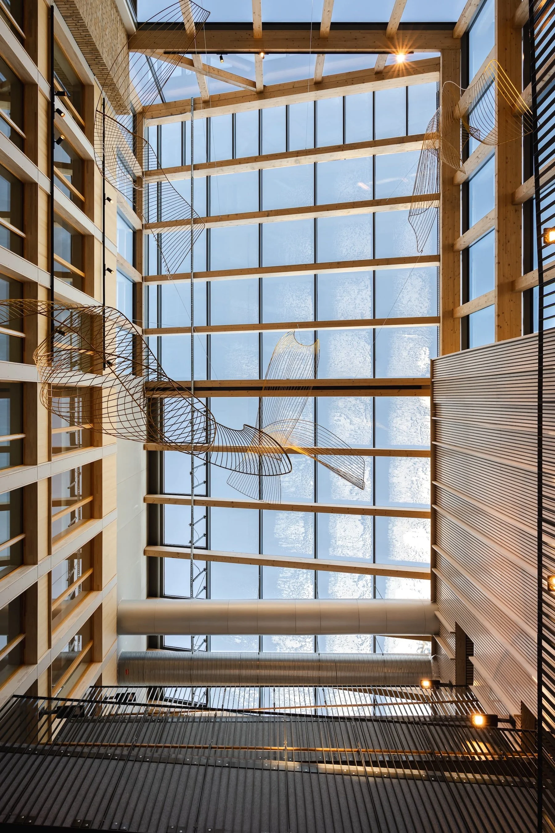 Indoor view of a modern building's atrium with large glass ceiling, wooden beams, metal artwork, and a wooden wall with black railings.