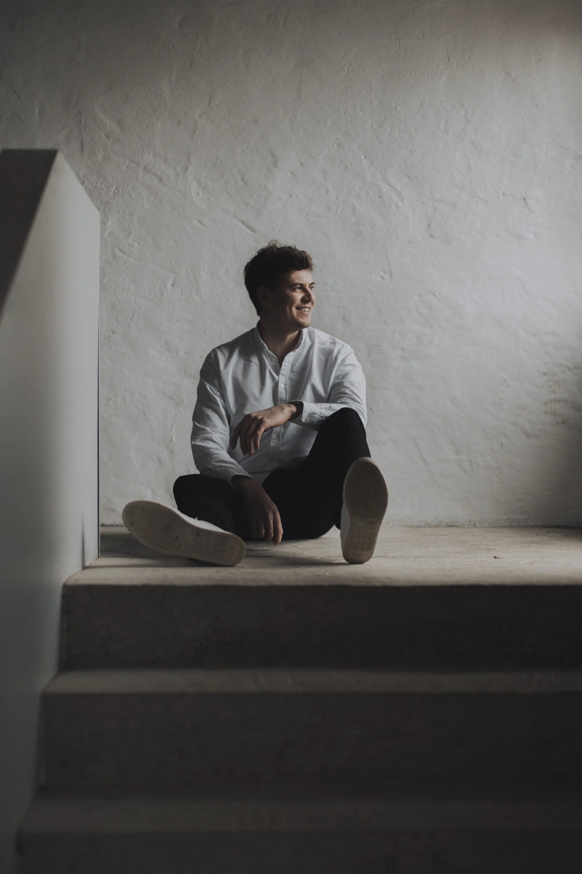 A young man with curly dark hair, wearing a white shirt and black pants, sitting on the carpeted floor at the top of a staircase, smiling and looking to his right, with a textured white wall behind him.