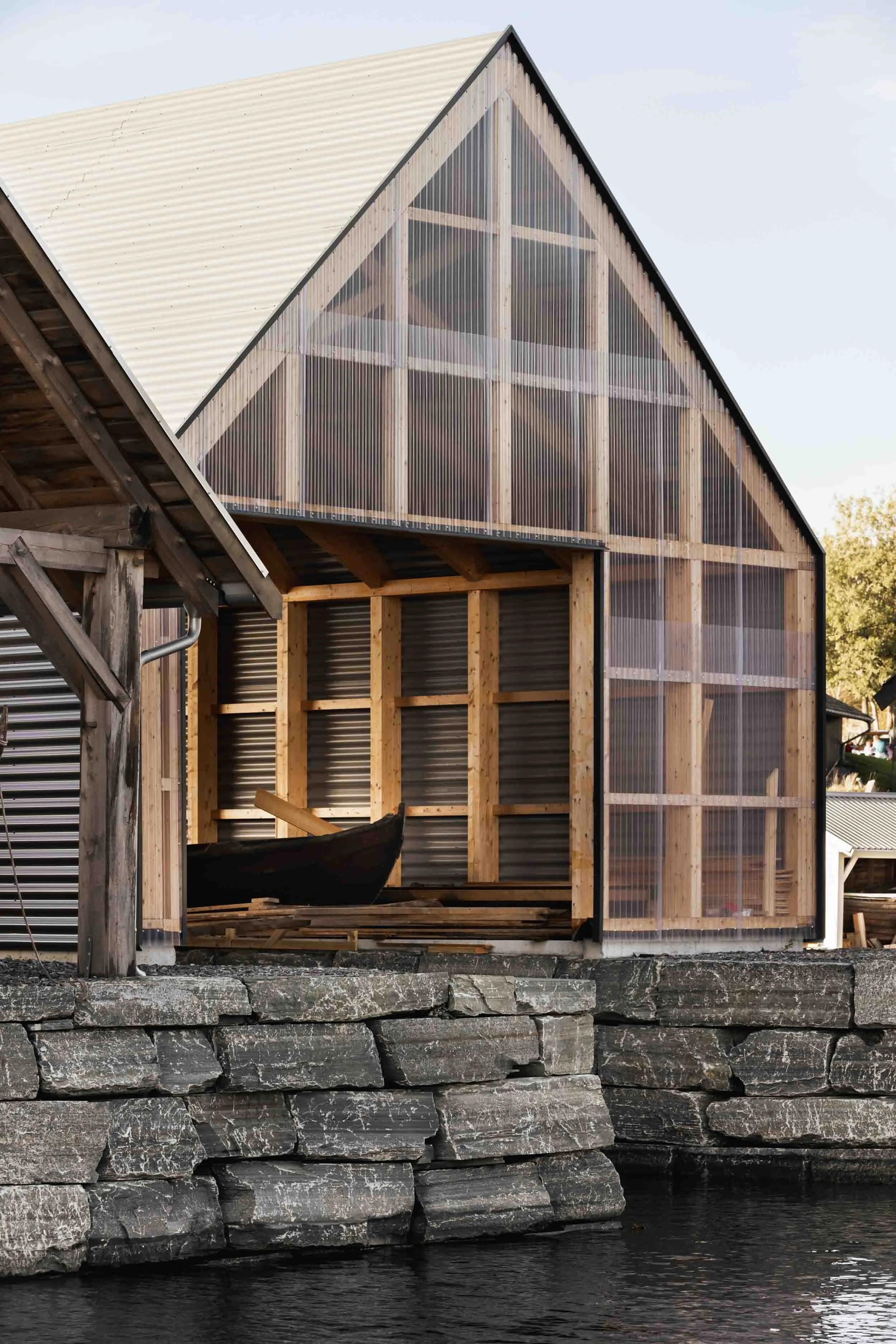 A building under construction with visible wooden framing and partially installed corrugated metal and plastic panels, situated on a stone foundation near water.