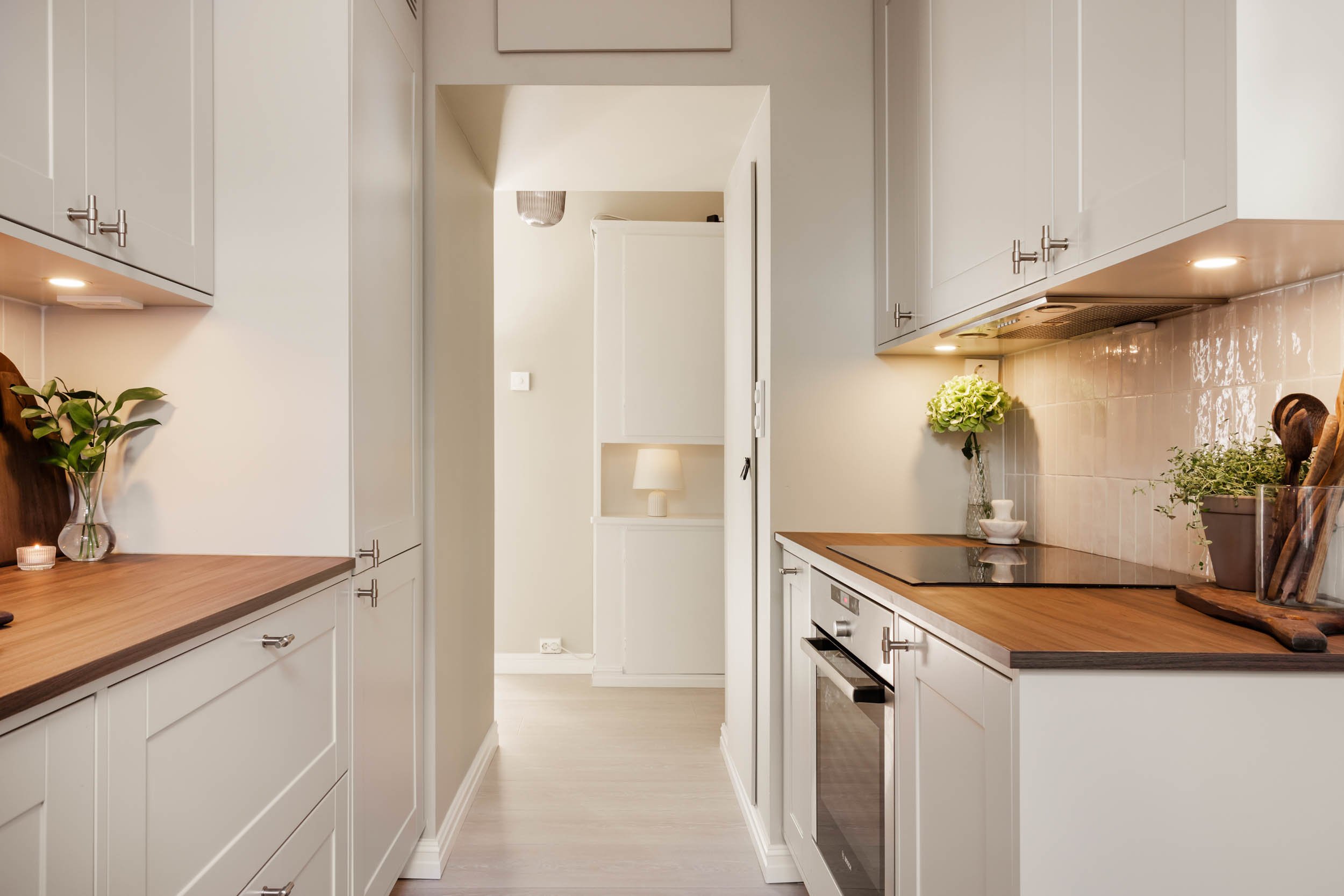 A modern kitchen with white cabinets, wooden countertops, and a minimalist decor, including plants, a lamp, and kitchen utensils.