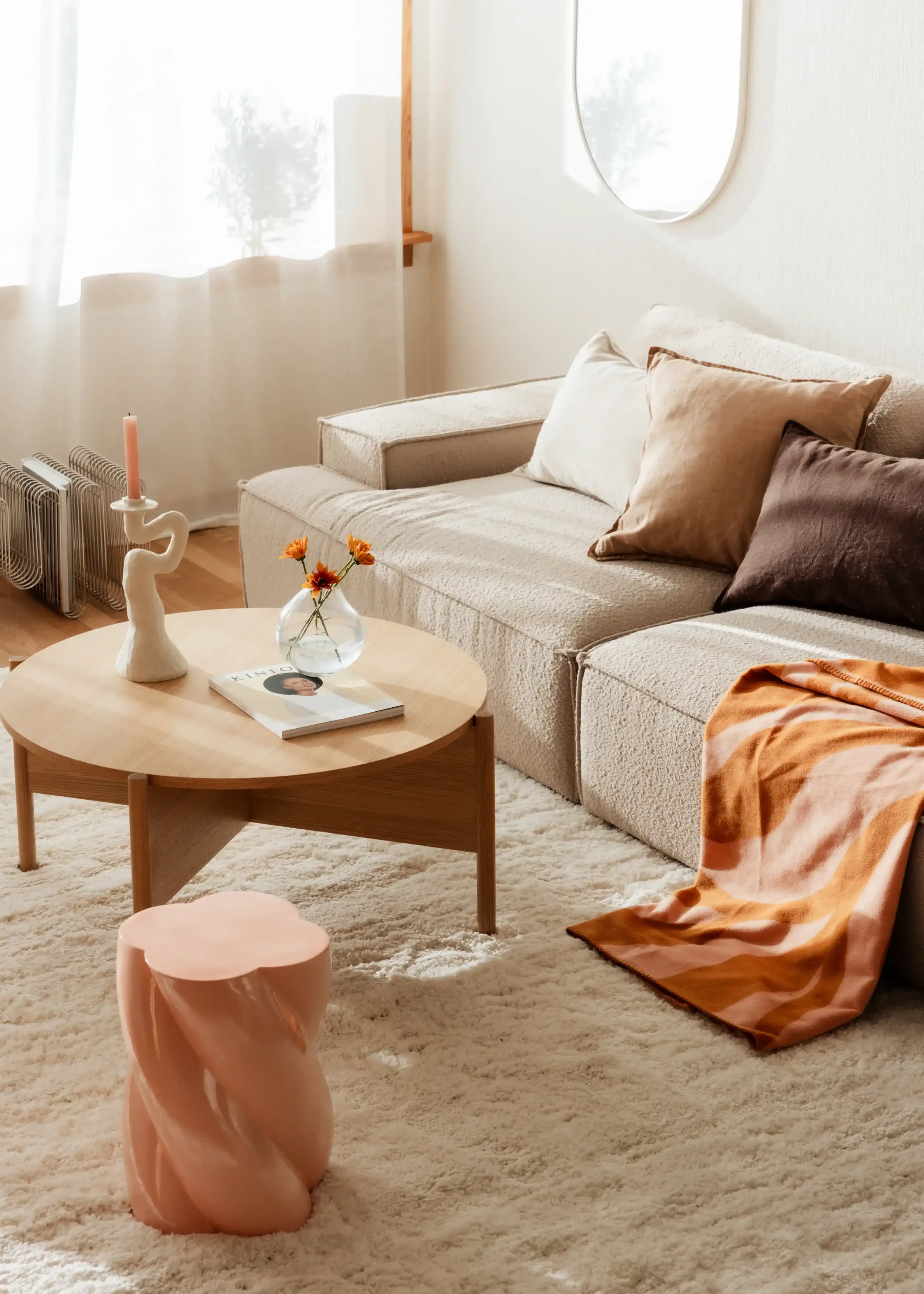 A cozy living room with a beige sofa, a round wooden coffee table with a flower vase and a magazine, a pink sculptural side table, a cream-colored fluffy rug, and a wall mirror. There are some cushions on the sofa, a throw blanket on the floor, and soft natural light coming through the window.