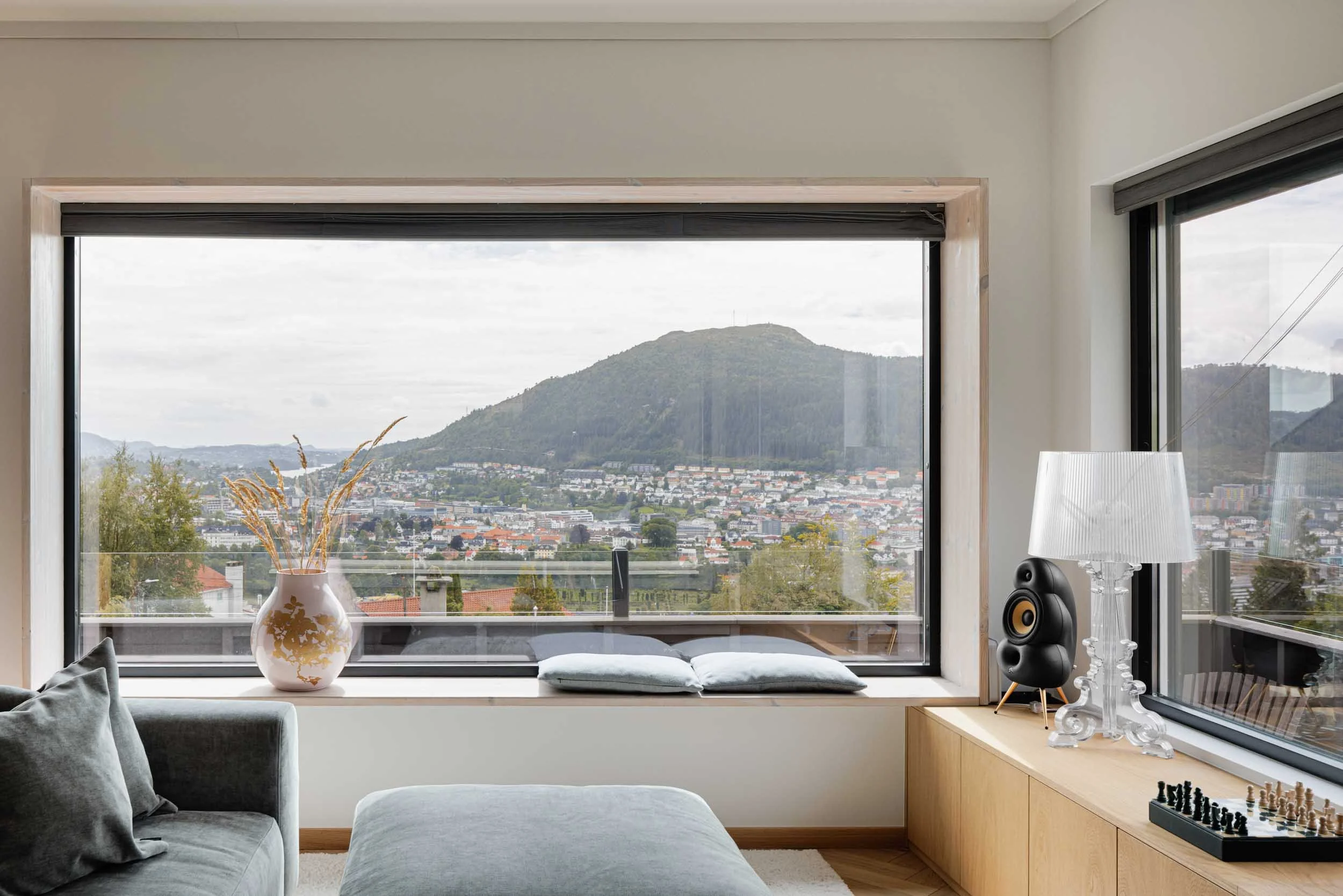 Living room interior with large windows showing cityscape and a mountain, a vase with dried plants on the window sill, a gray sofa with pillows, a side table with a chessboard, a clear glass table lamp, and a black speaker. Fjellsiden, Bergen