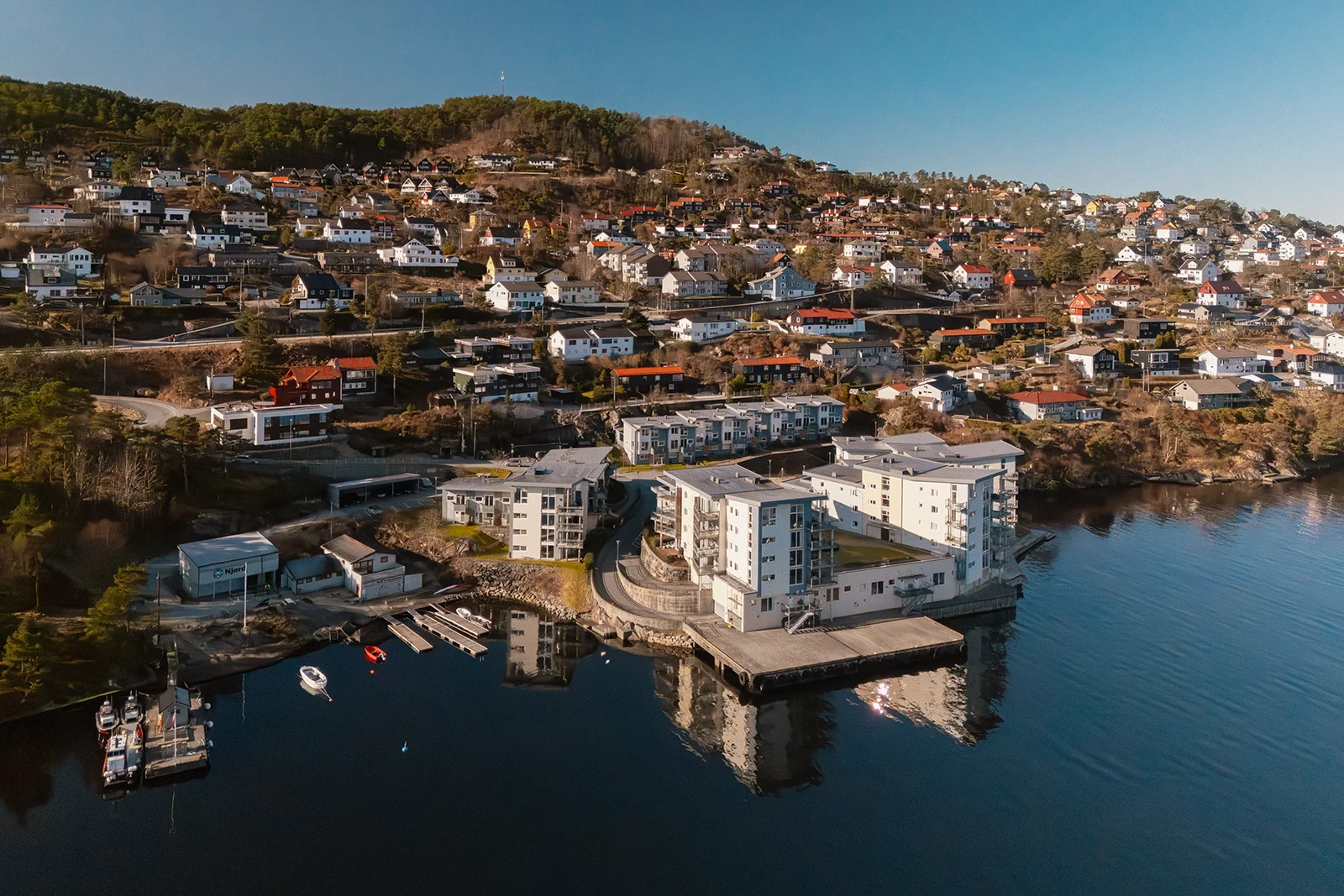 Aerial view of a hillside residential area with houses and an apartment complex by the water, with boats docked at the marina