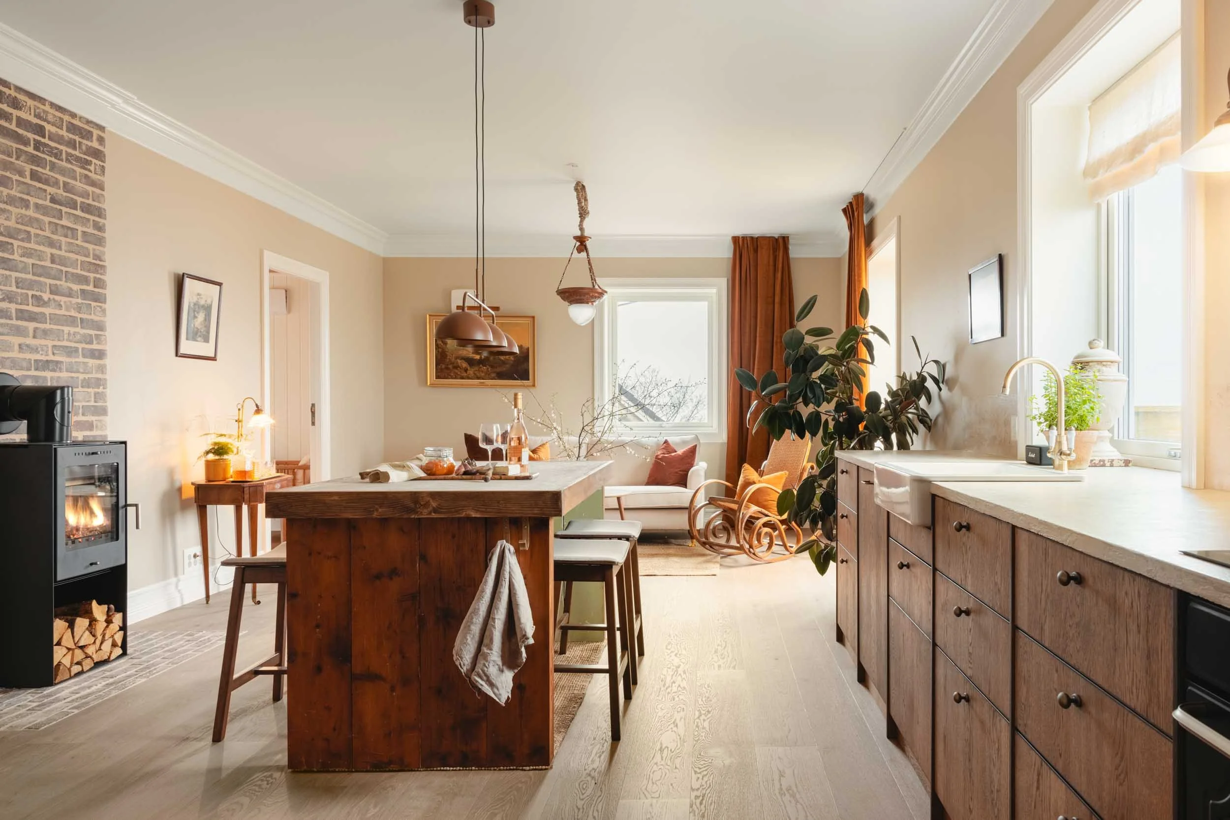 Open-concept kitchen and living room with wooden island, light wood floors, brick and beige walls, natural light from large windows, potted plant, rocking chairs, and minimalist decor.