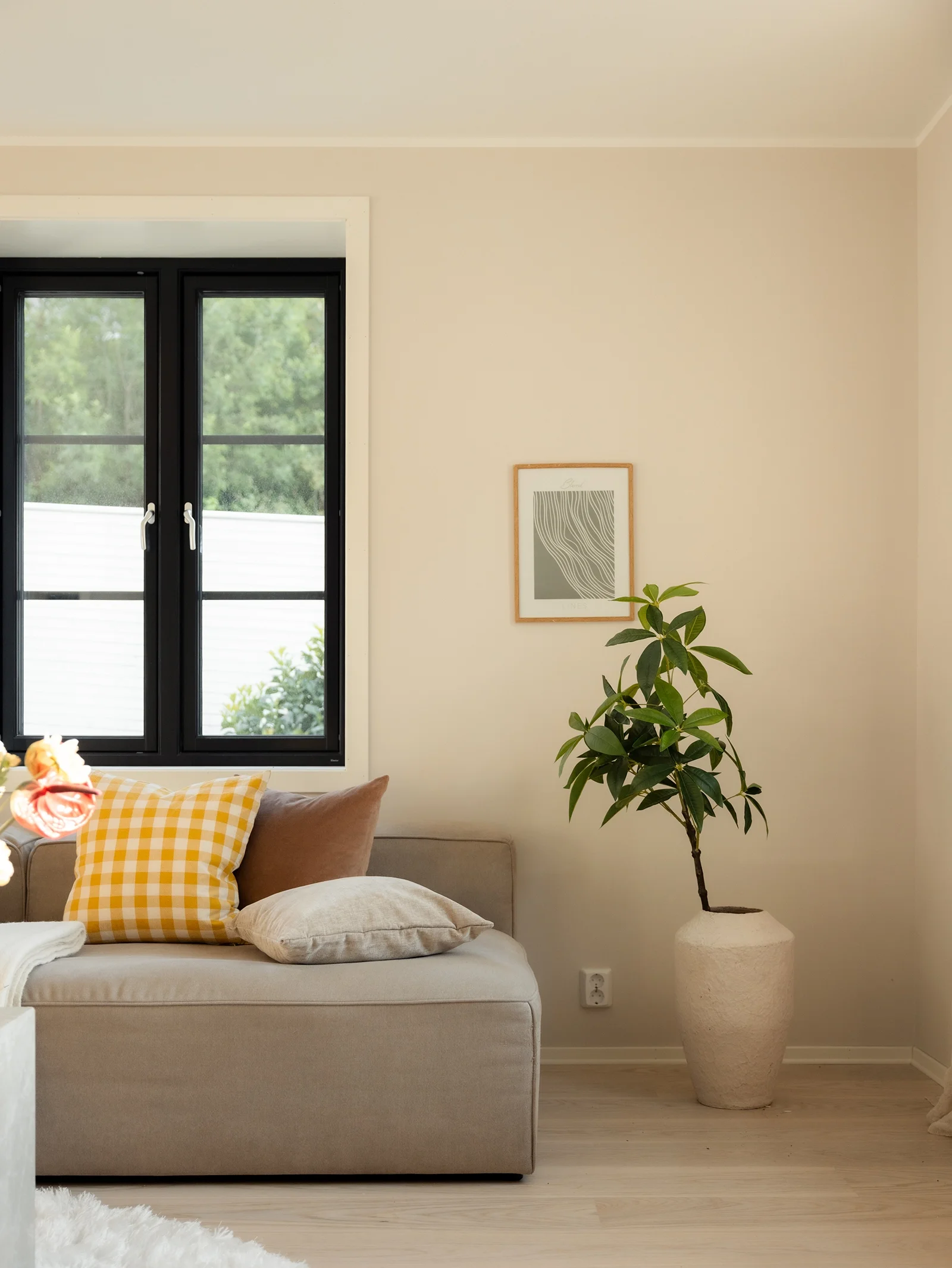 Living room with a beige sofa, yellow and checkered pillows, a potted plant, and a framed artwork on the wall.