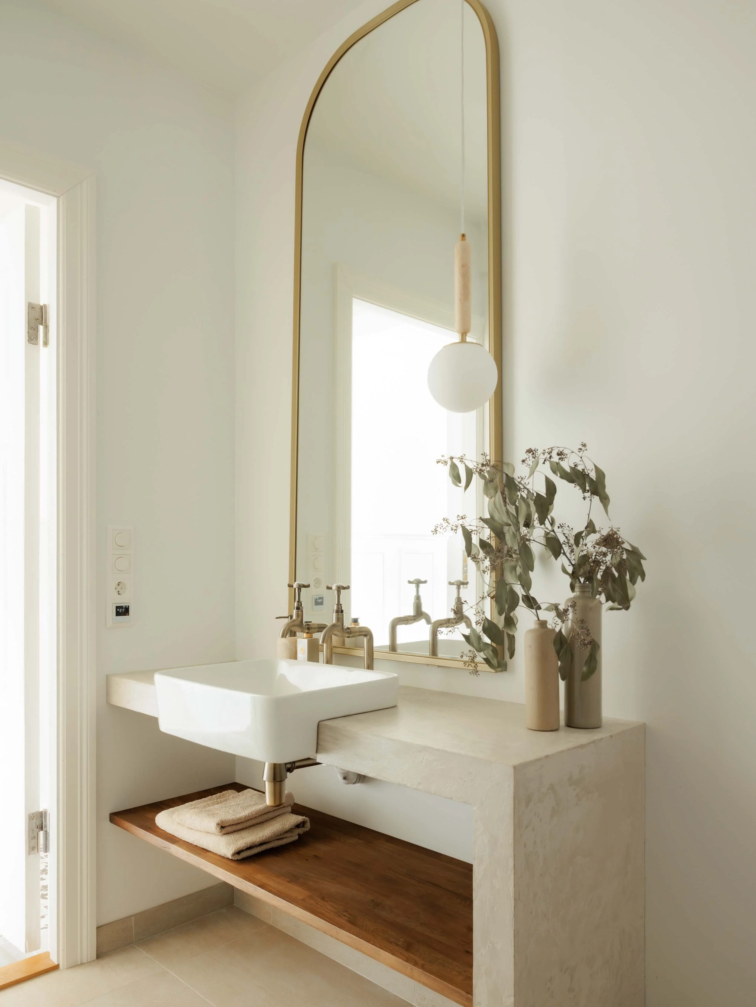Minimalist bathroom with a rectangular white sink, a large gold-framed mirror, two small vases with dried leaves, a hanging light with a round white globe, and a wooden shelf with folded towels.