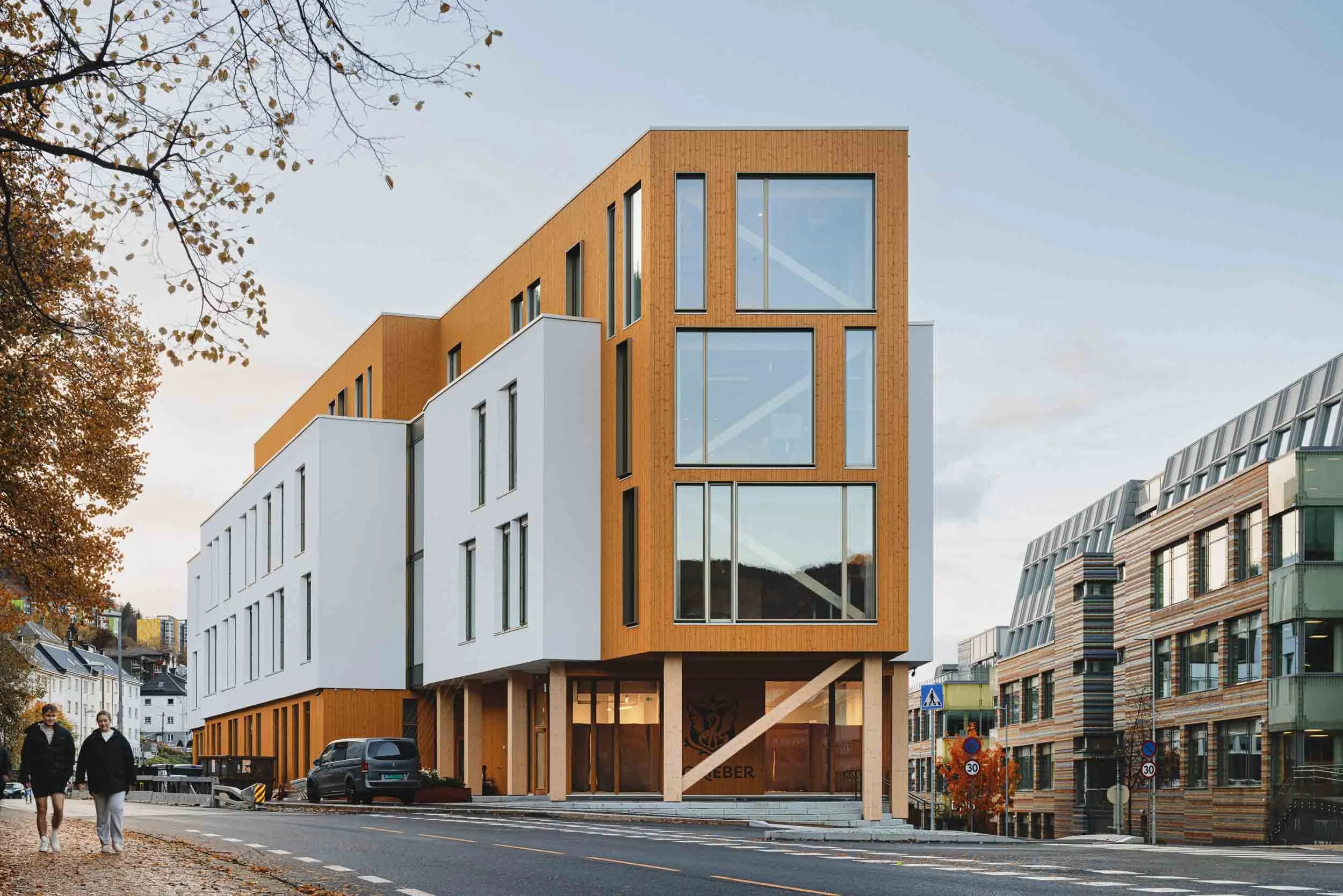 Modern multi-story building with wooden and white facade, large windows, situated on a city street. Two people walking on the sidewalk in fall, trees with autumn leaves.