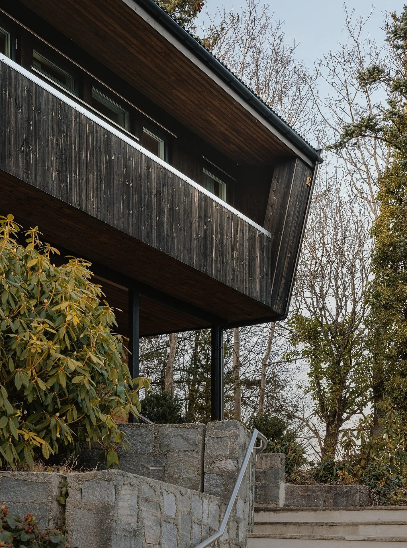 A modern house with a slanted dark wooden exterior, situated on a stone staircase surrounded by bushes and trees, during early spring. - Askøy