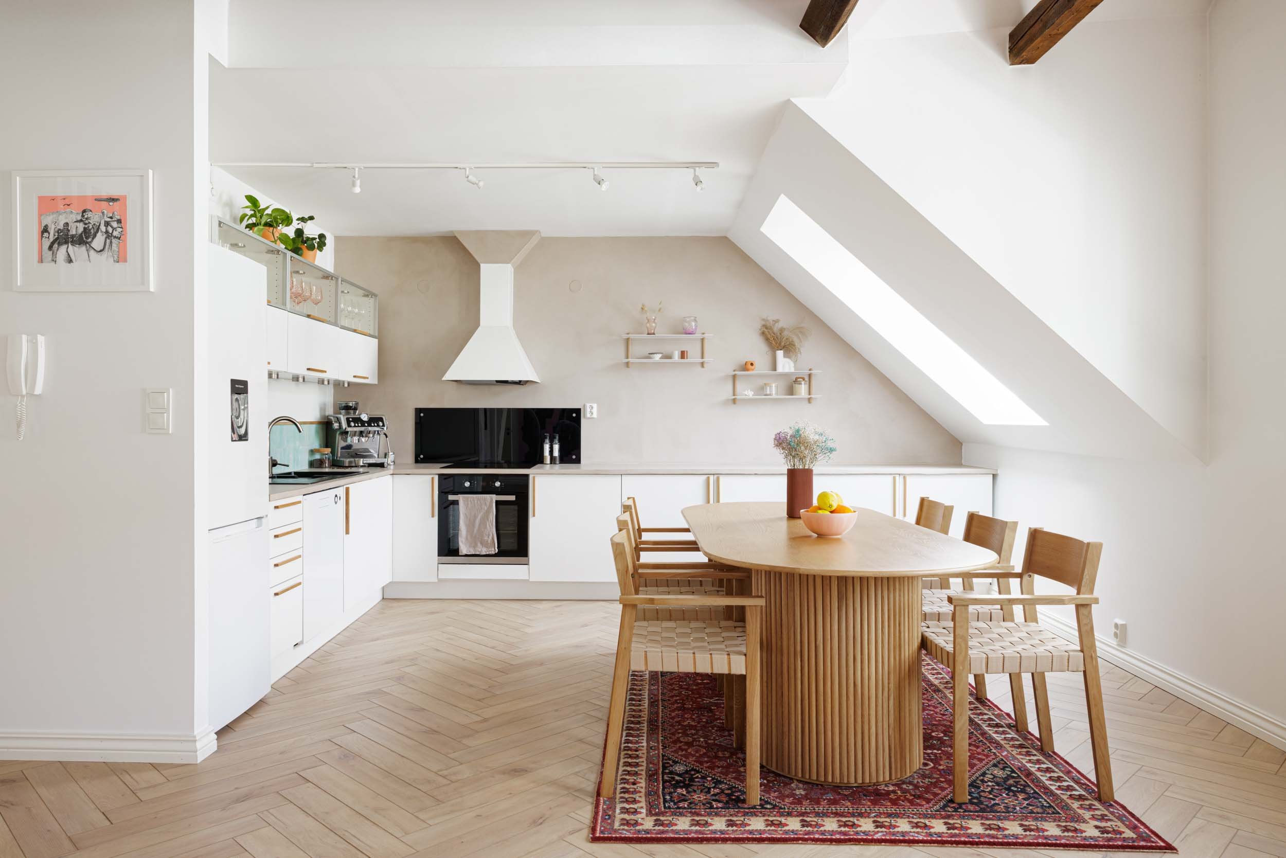 Modern kitchen and dining area with white cabinets, black appliances, and a wooden dining table with chairs on a red patterned rug, featuring a skylight and decorative shelves.