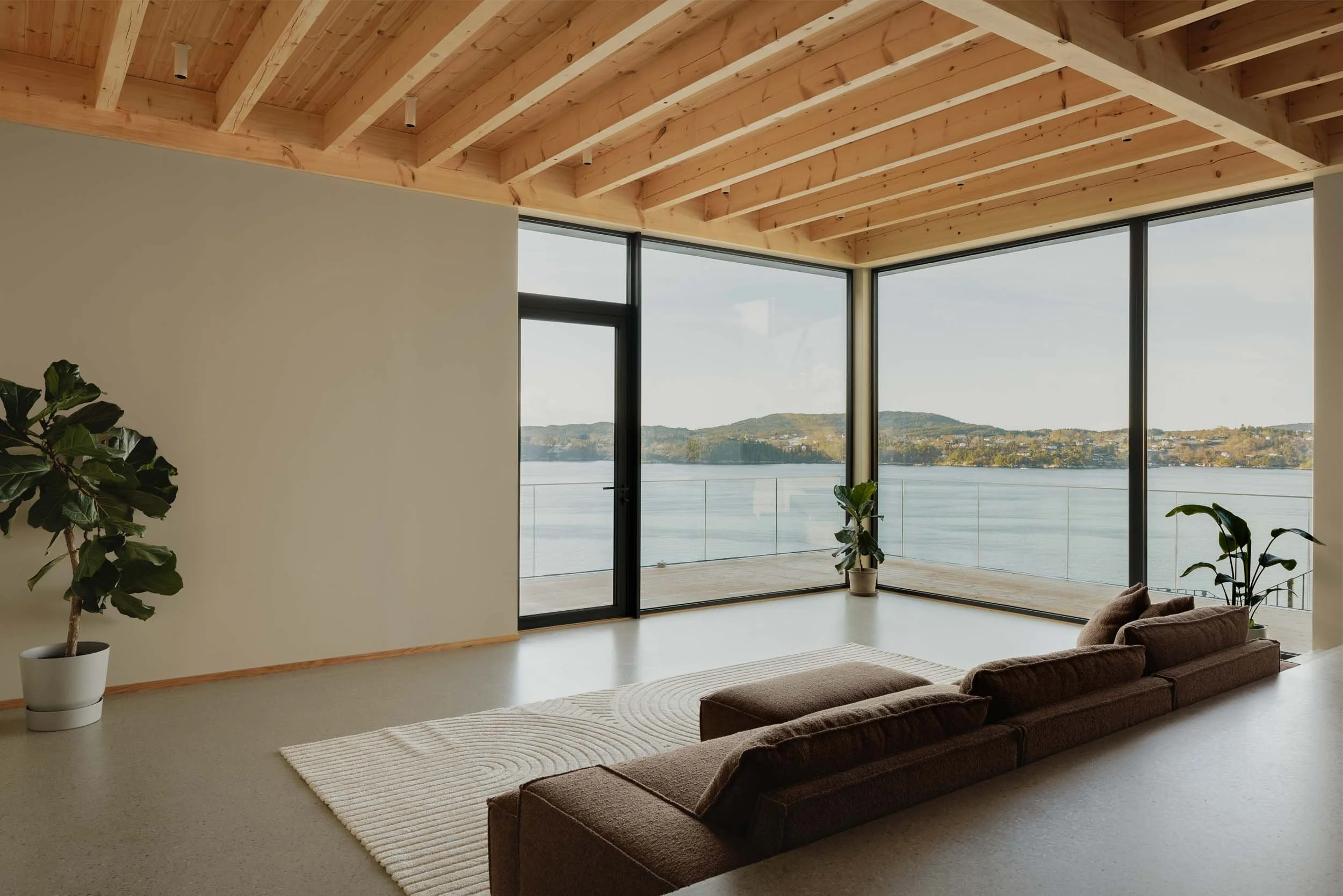 Modern living room with large floor-to-ceiling windows overlooking a body of water and distant hills, wooden ceiling beams, beige walls, brown sectional sofa, potted plants, cream rug.