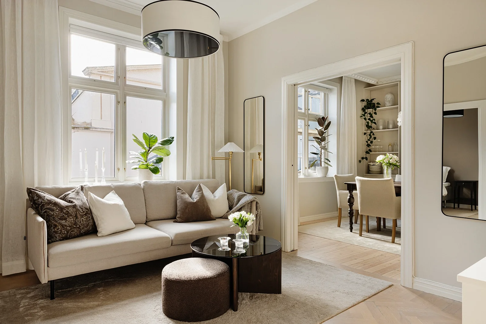 Living room with beige sofa, white curtains, green plants, black and glass coffee table, and decorative pillows. Adjacent dining area with a table and chairs, decorated with flowers and plants.