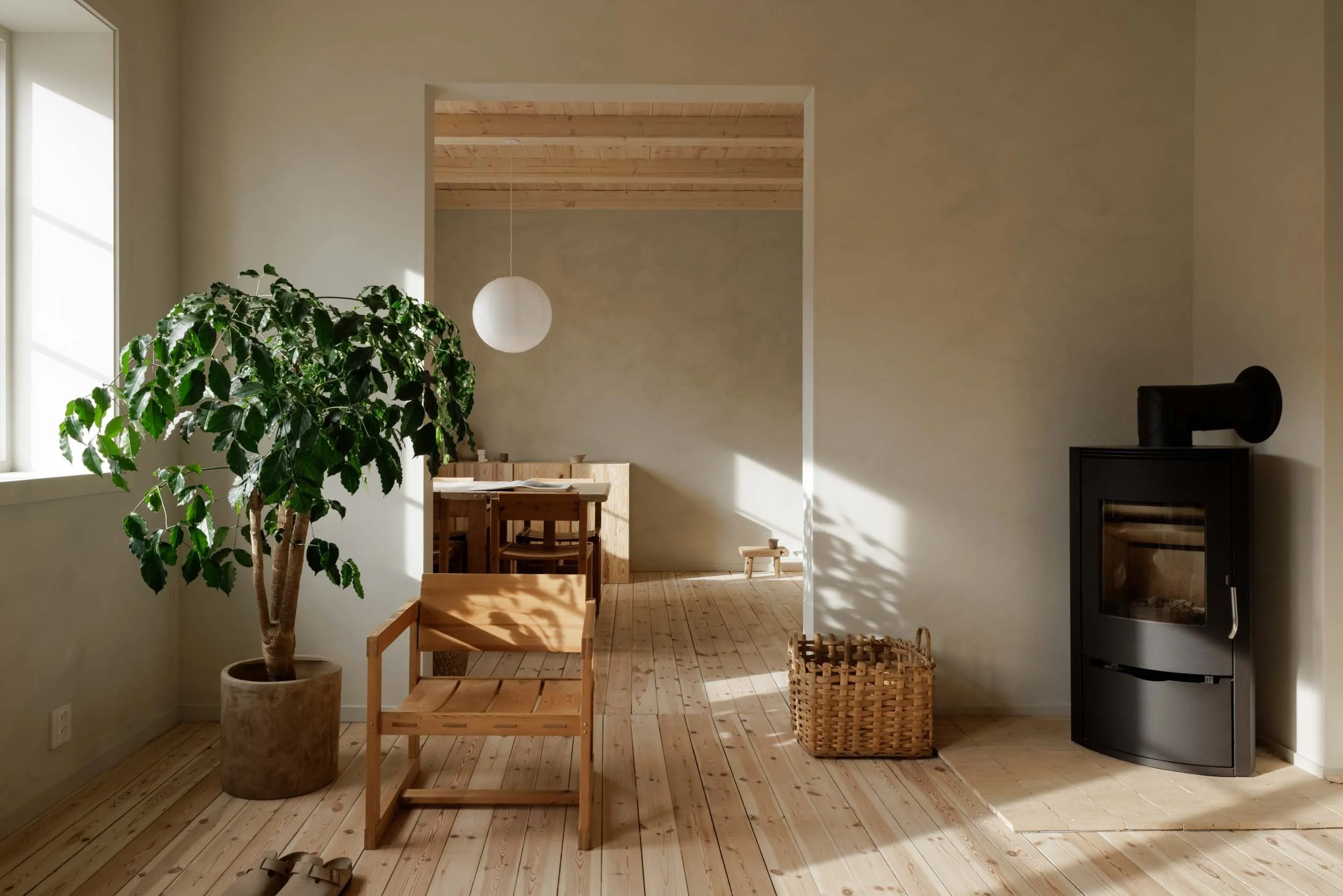 Living room with wooden flooring, a green potted plant, a wooden chair, a basket, a wood stove, and sunlight streaming through a window.