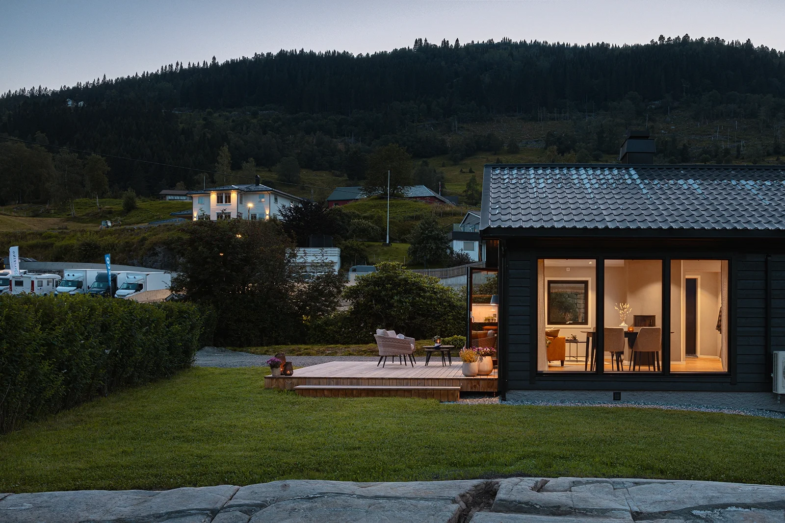 View of a house with large windows and a patio with outdoor furniture, on a grassy lawn at dusk, with a wooded hillside and other houses in the background.