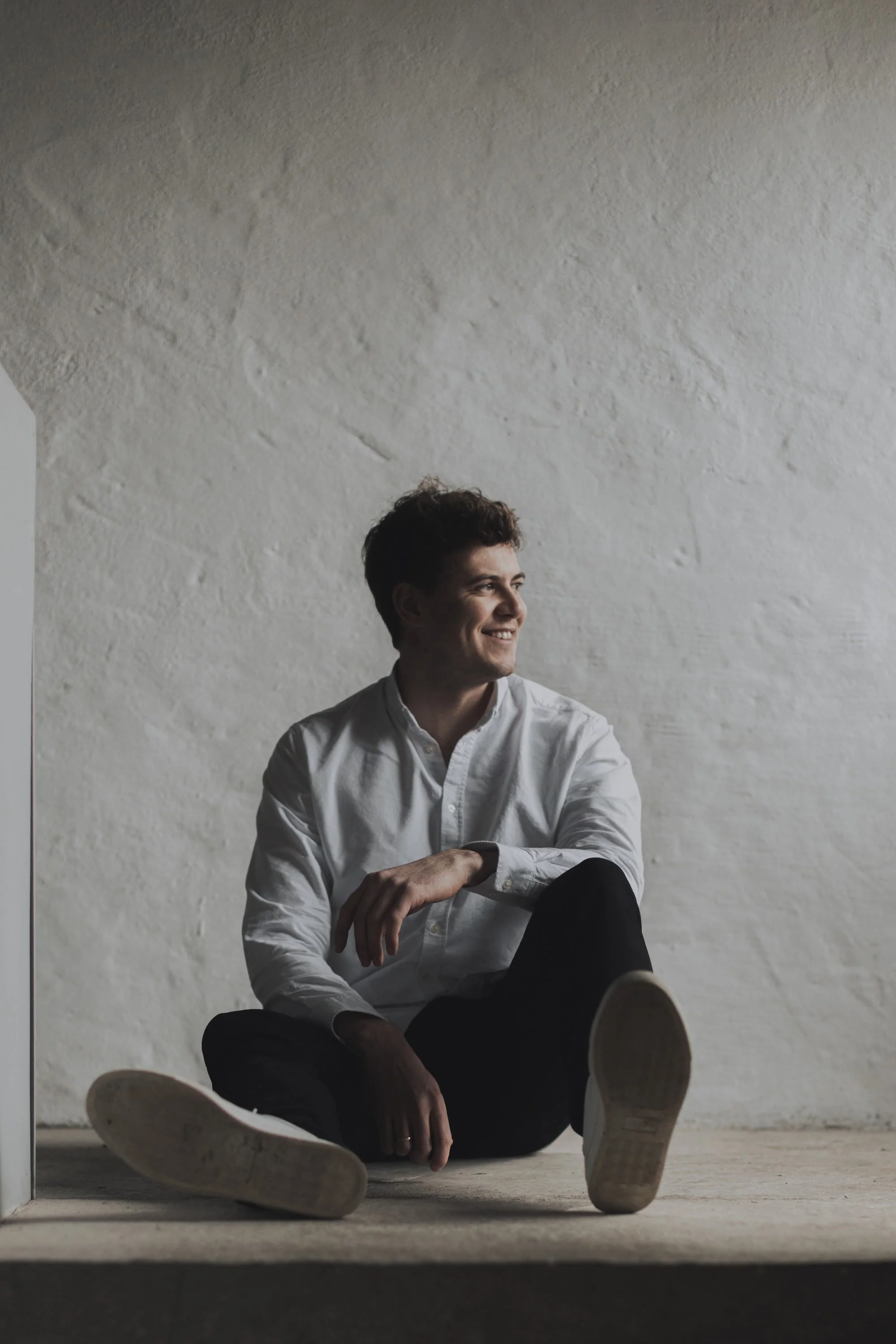 A young man with dark curly hair dressed in a white shirt and black pants sitting on a raw concrete floor against a concrete textured wall, smiling and looking to his right.