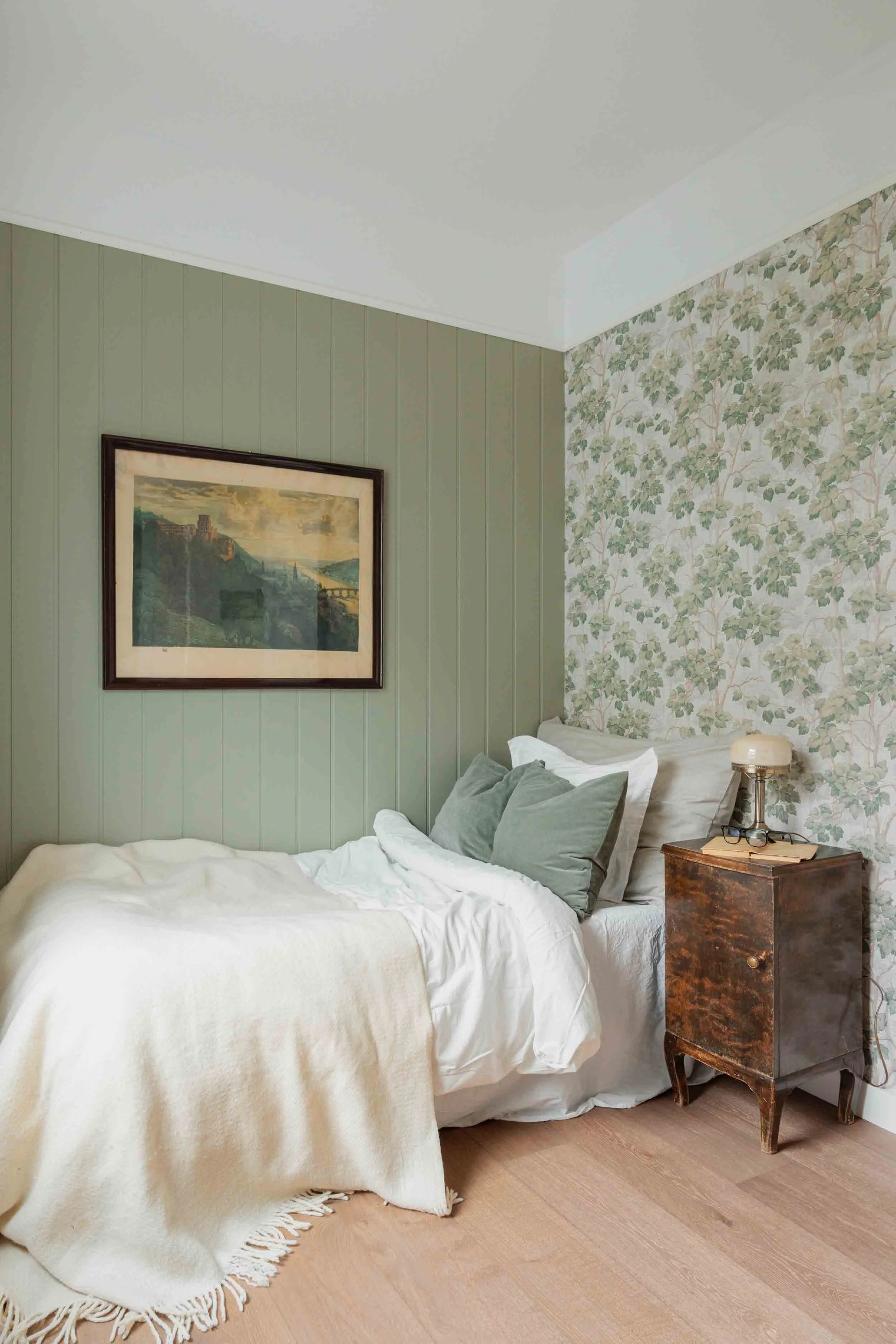 A cozy bedroom with a bed covered in white linens and gray pillows, a vintage wooden nightstand with a lamp and glasses, framed artwork on a green panel wall, and floral wallpaper on the adjacent wall.