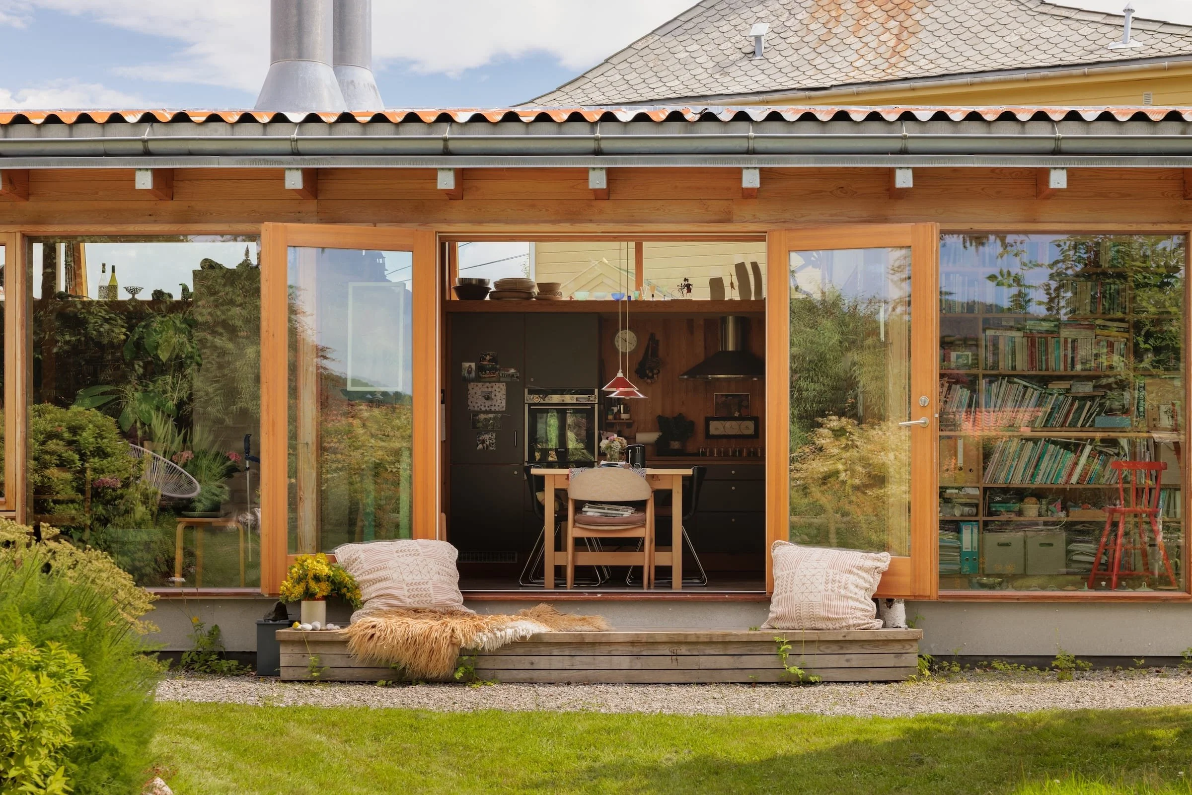 The back view of a modern house with large glass sliding doors opening to a garden. Inside, a kitchen and dining area are visible, featuring cabinets, a black refrigerator, a wooden dining table with chairs, and a hanging red pendant light. Outside, there are cushions and a fur throw on a wooden step with a small flowerpot on the left.