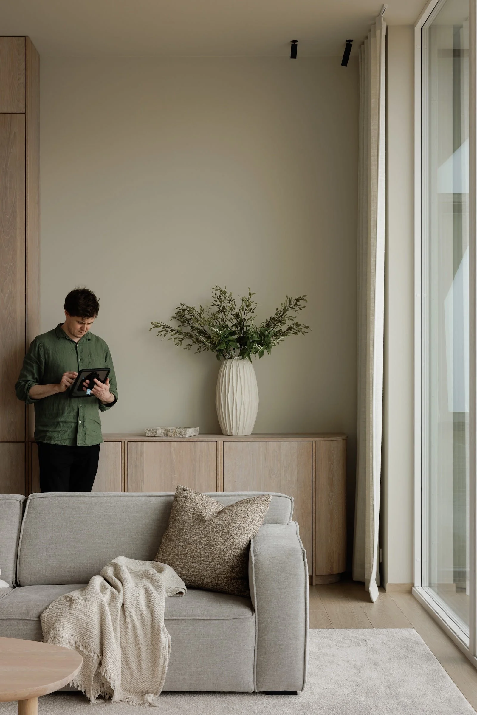 A man standing in a living room using a tablet with a beige sofa with textured pillows and a blanket in front of him, a wooden cabinet with a vase of greenery and a stone on top, large windows letting in natural light, and neutral-colored decor.