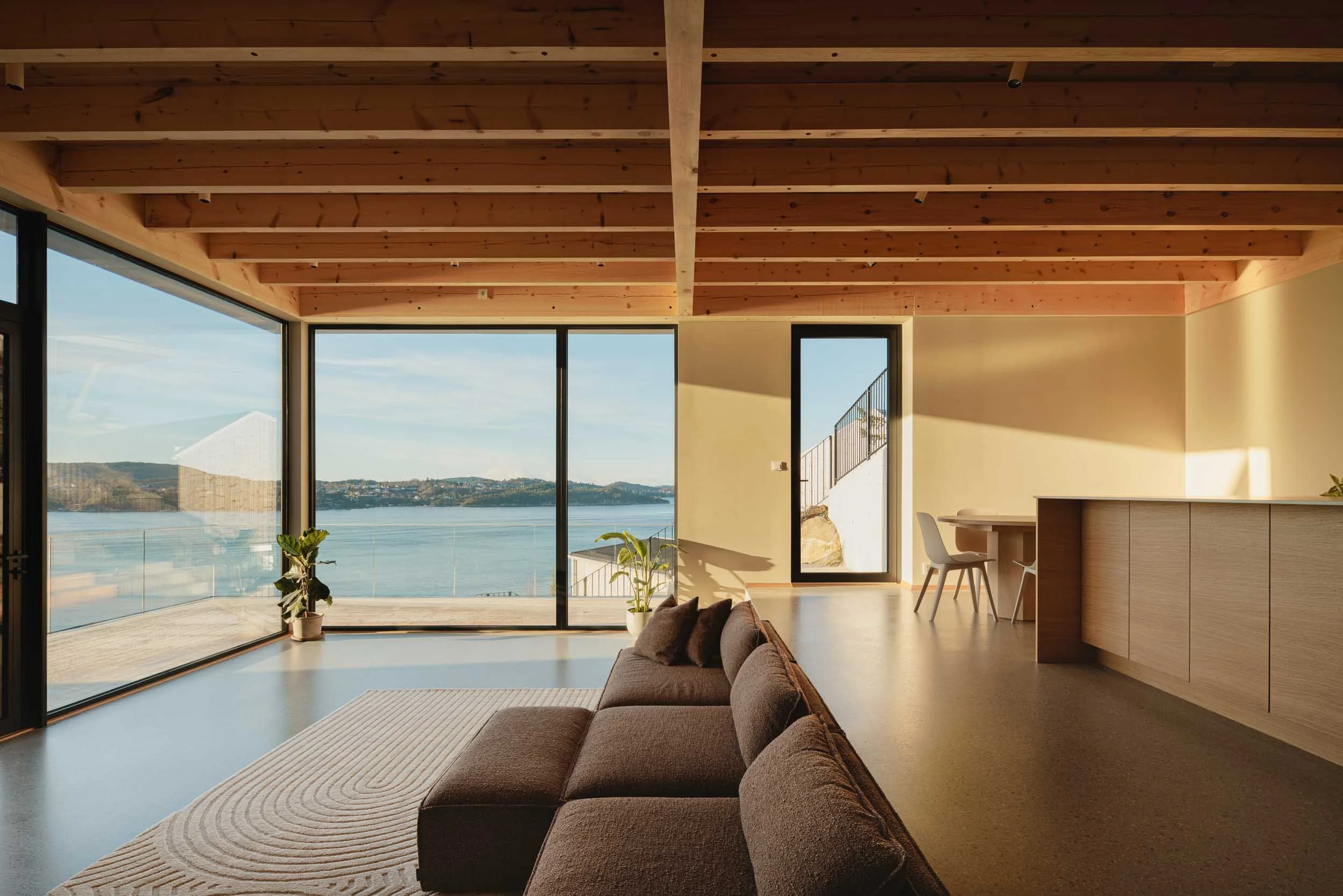 Living room with large glass windows overlooking a body of water, modern furniture, and wooden ceiling beams.