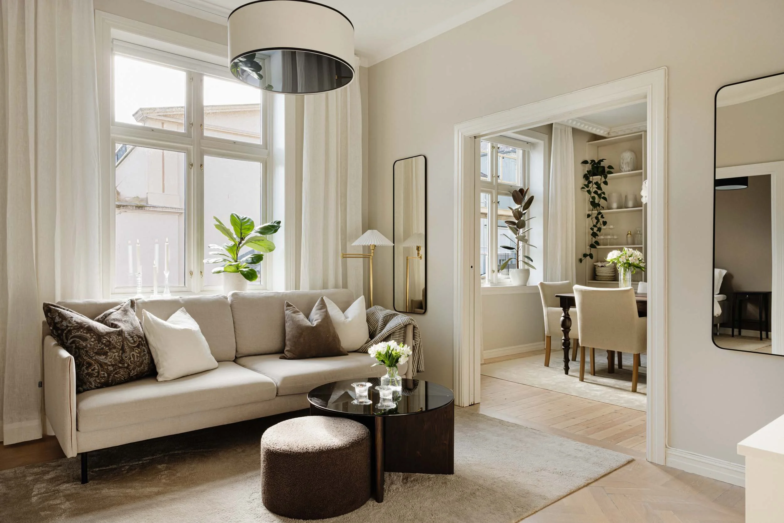 A bright living room with large windows, a beige sofa with decorative pillows, a black round coffee table with a small vase of flowers, a large ceiling light, and a mirror on the wall. There is an adjoining dining area with a table, chairs, and decor in the background.