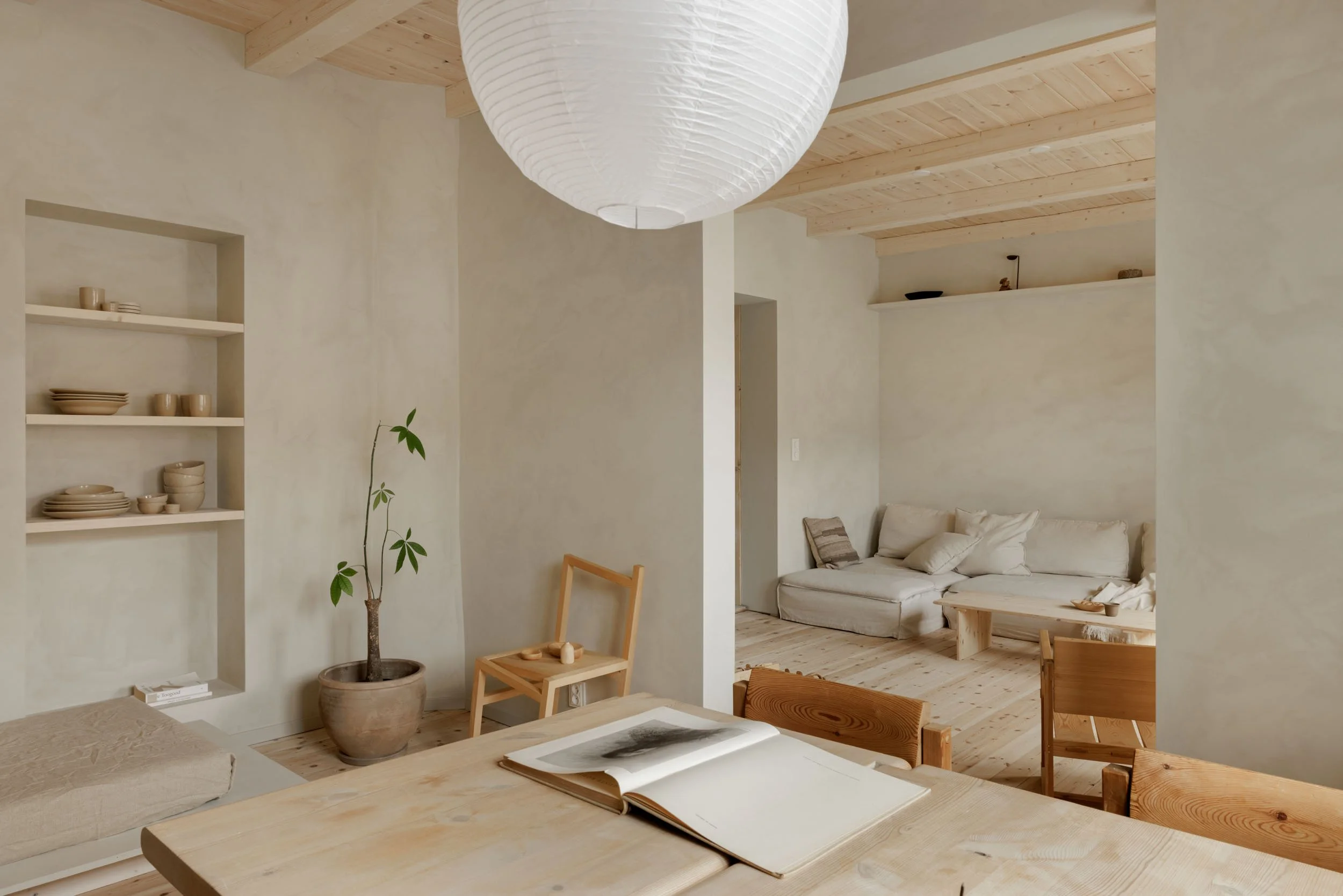 A cozy living room with minimalist decor, featuring a white couch with cushions, a wooden coffee table, a potted plant, and built-in shelves with dishes, all in neutral tones.