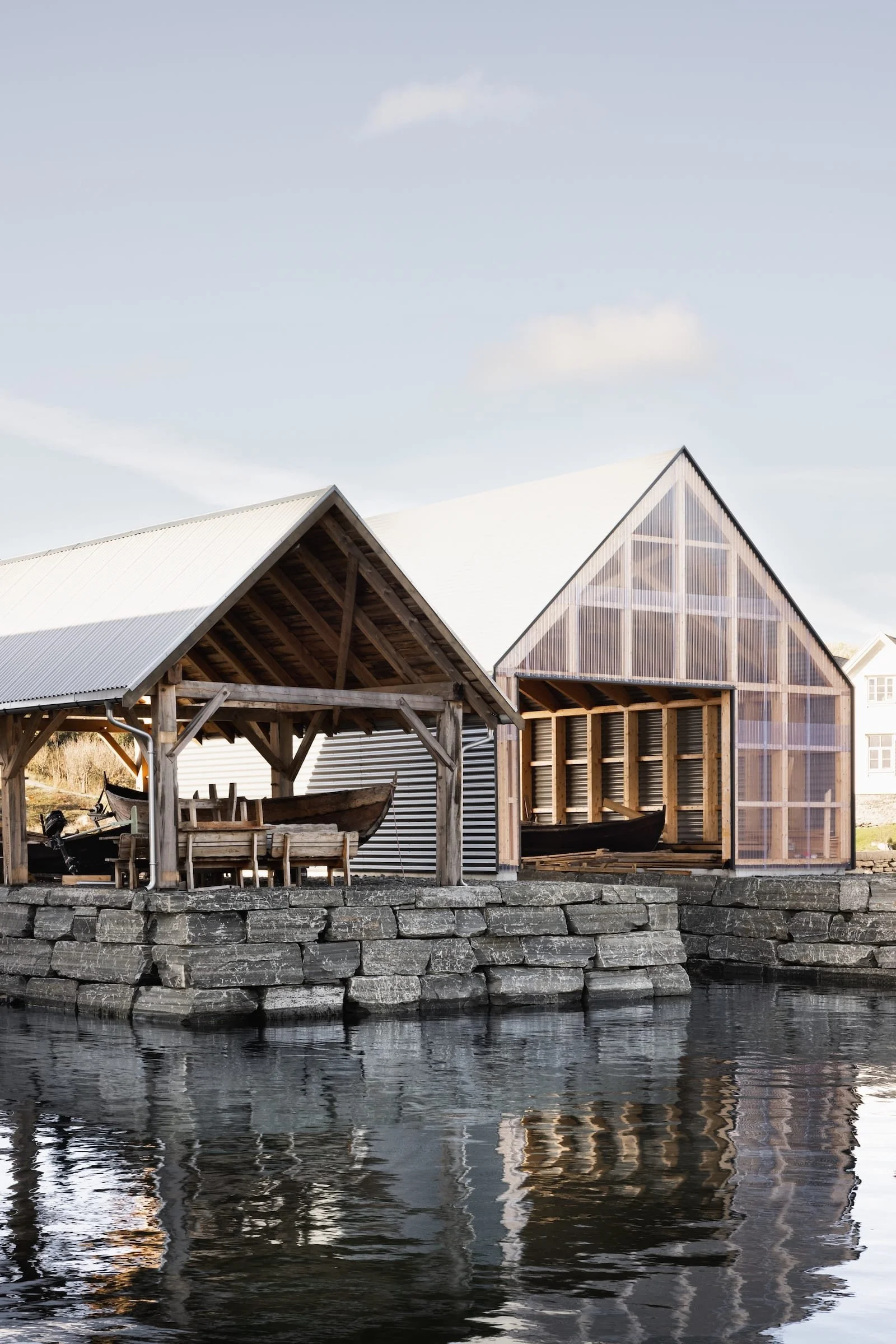 Two wooden boathouses with boats inside, located on a stone pier by the water under a light sky.