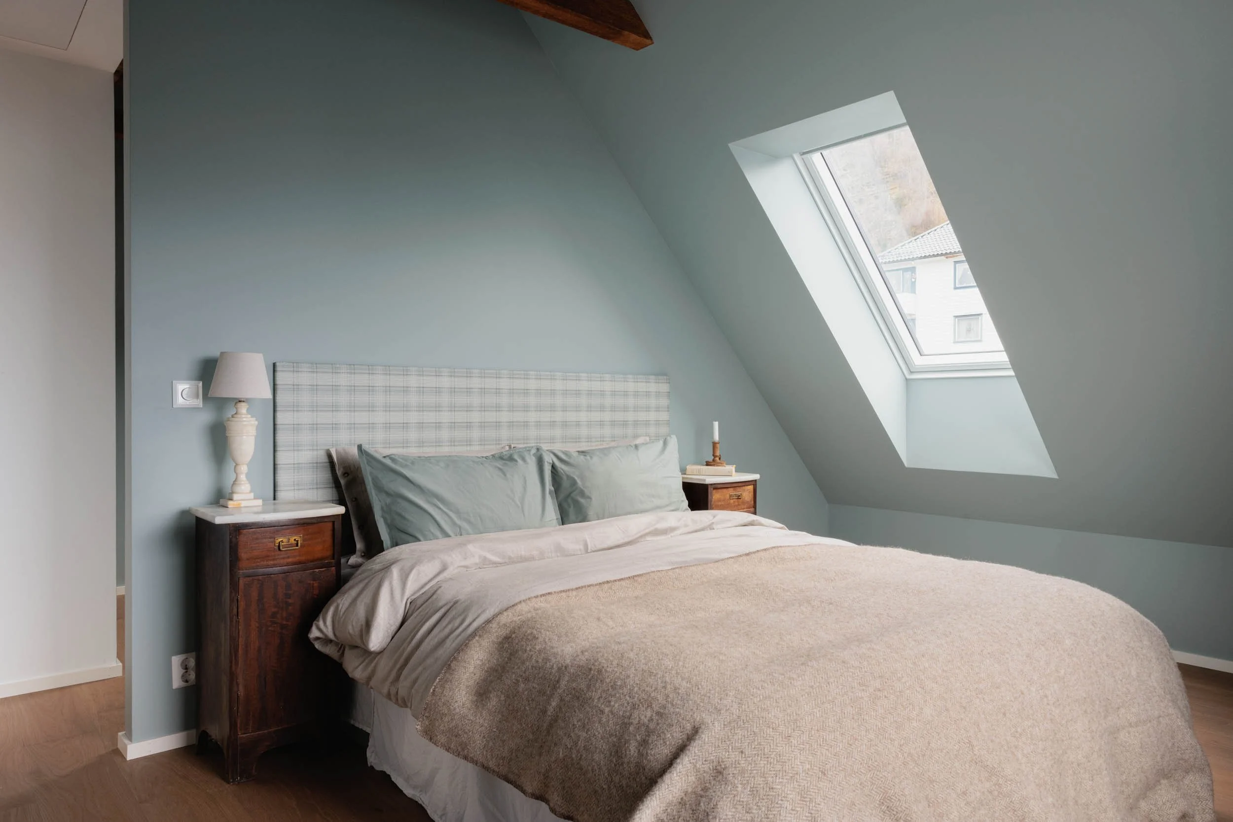 A cozy bedroom with a vaulted ceiling, featuring a bed with light grey pillows and a beige blanket, a retro wooden nightstand with a white lamp, and a large skylight window showing neighbouring buildings.