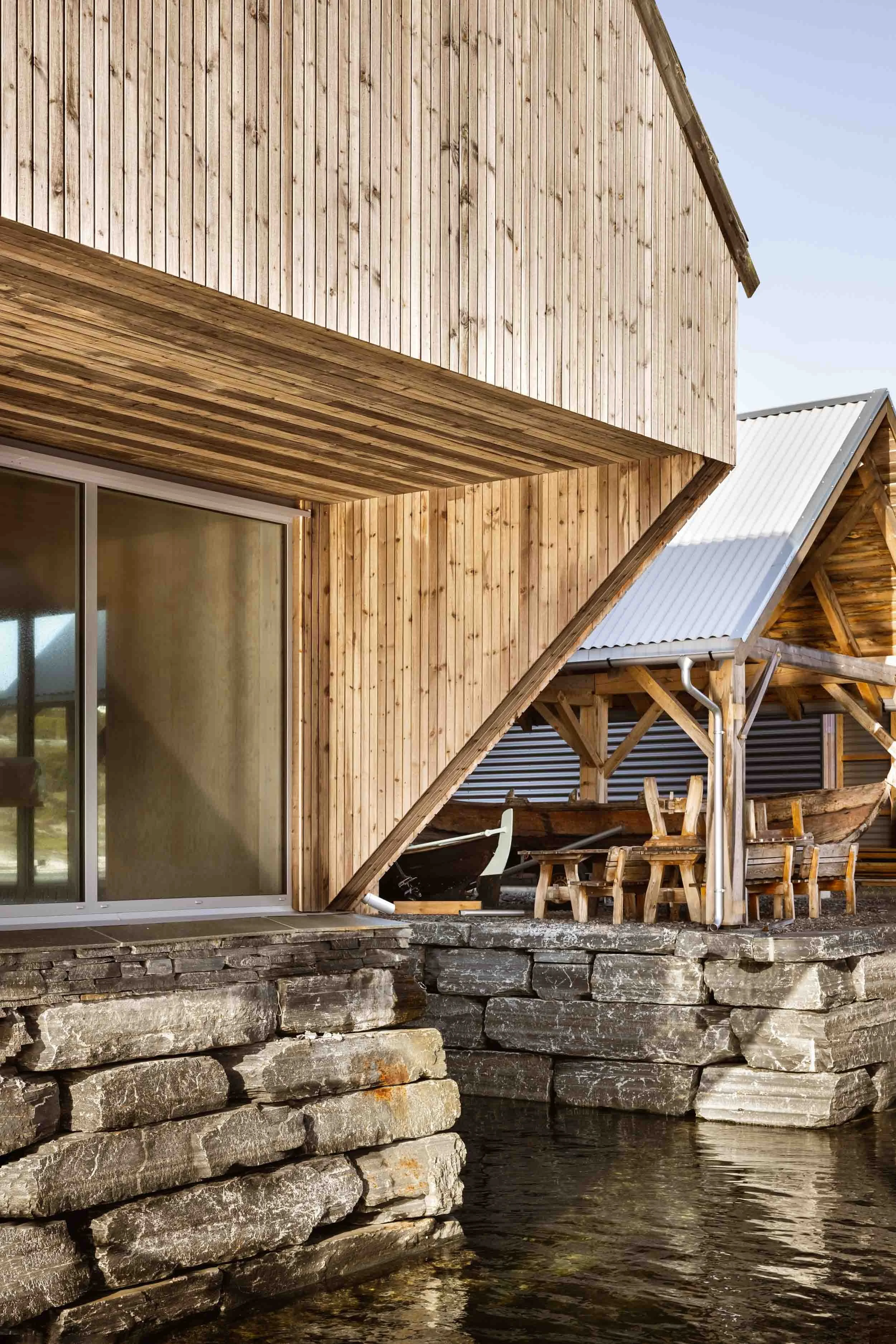 Modern wooden boathouse with large glass windows on a stone foundation by water, with boats and outdoor seating area in the background. Kystmuseet i Øygården