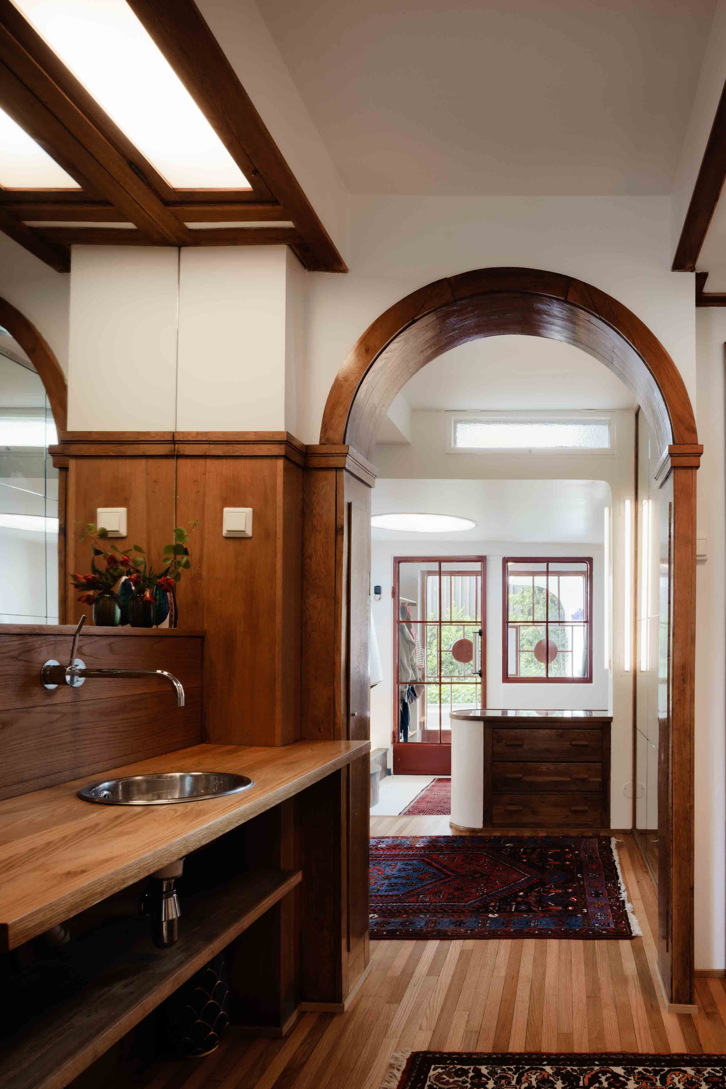 Indoor view of a home with wooden door and window frames, arched doorway, wooden flooring, and a hallway with a red and blue patterned rug.